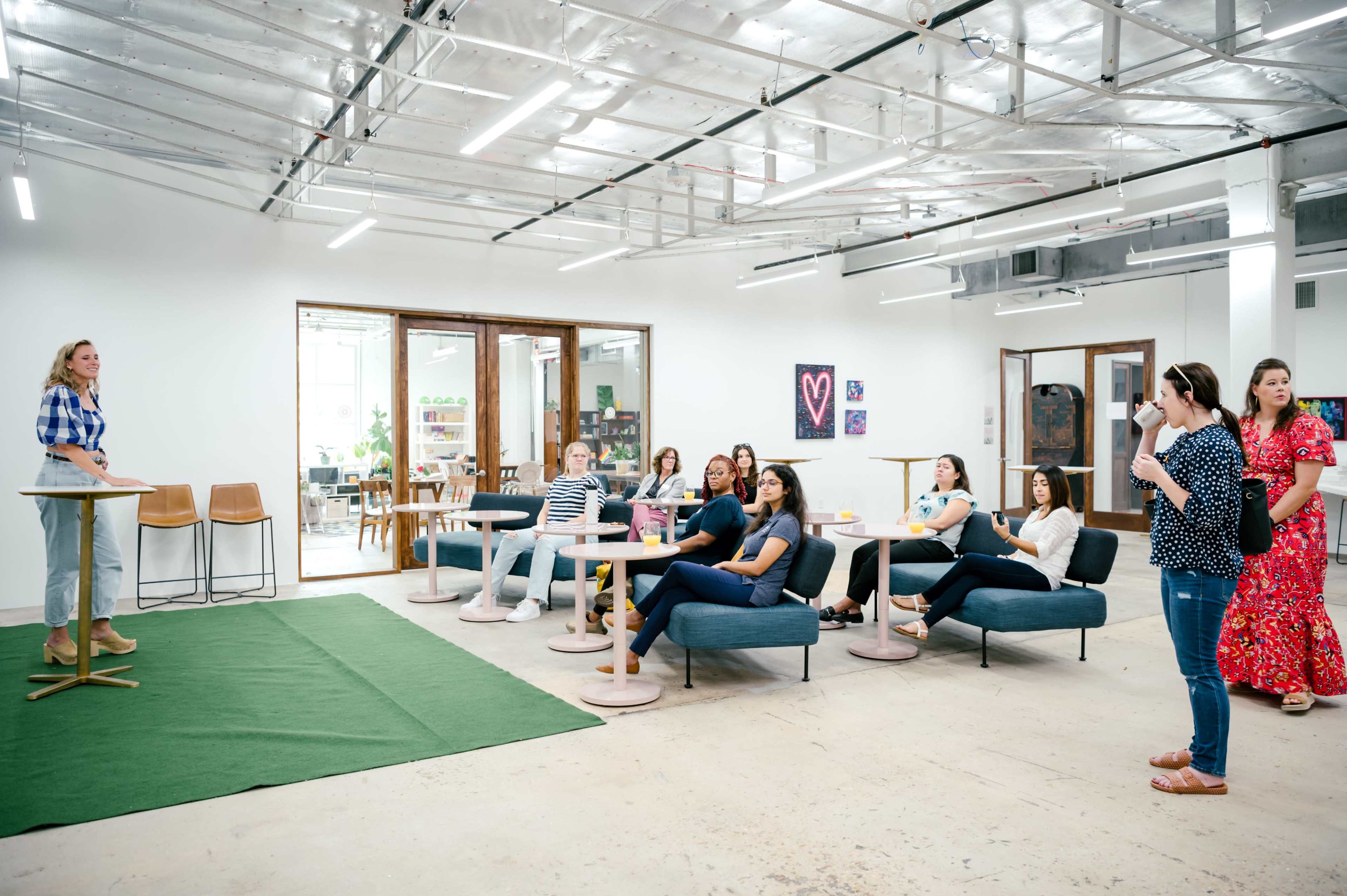 A group of people sits on couches and chairs in a bright, open space while another person stands at a small table.