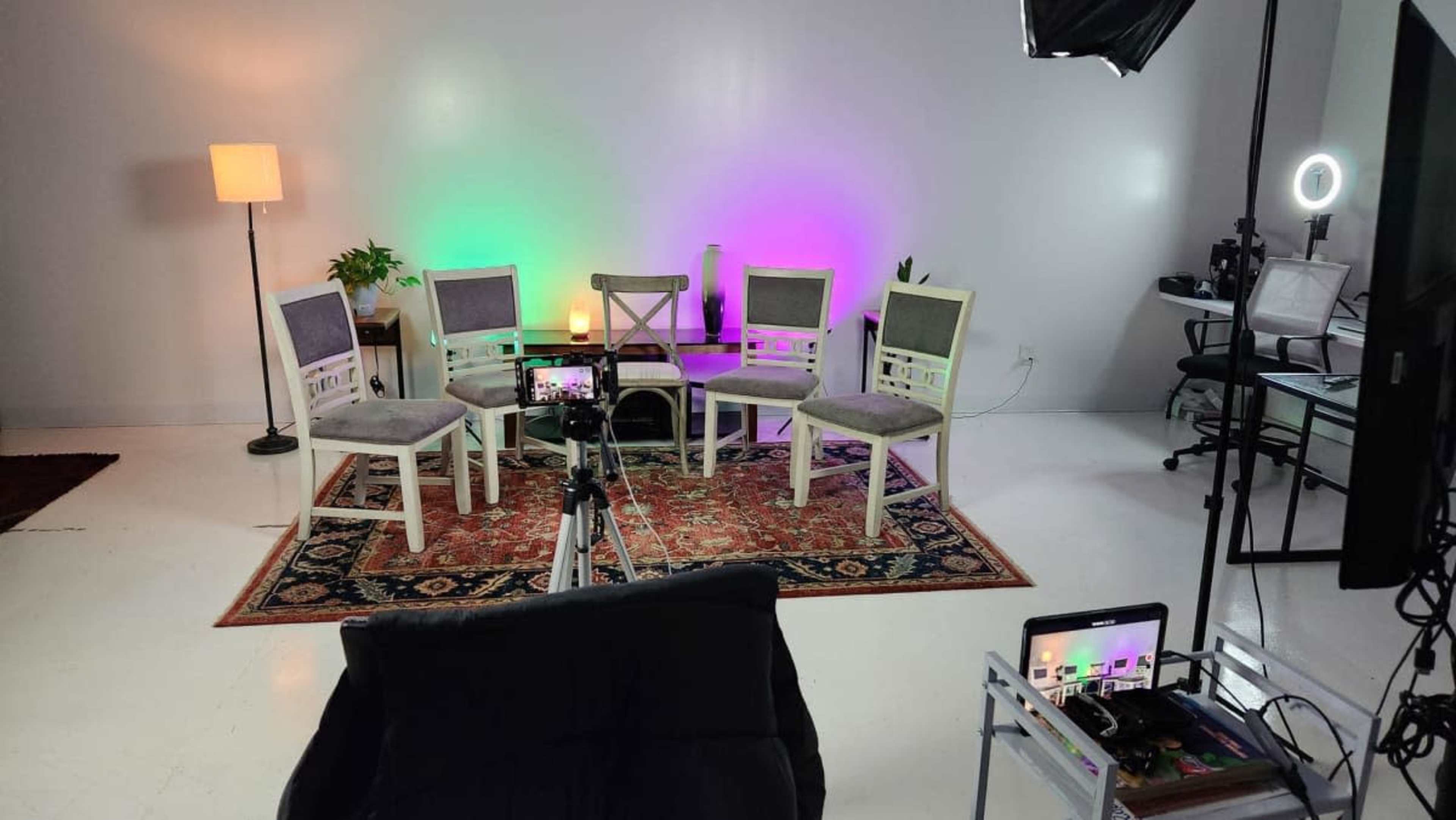 The image shows a setup for a video or photography shoot with four chairs arranged around a table, enhanced by colorful lights and a camera on a tripod.