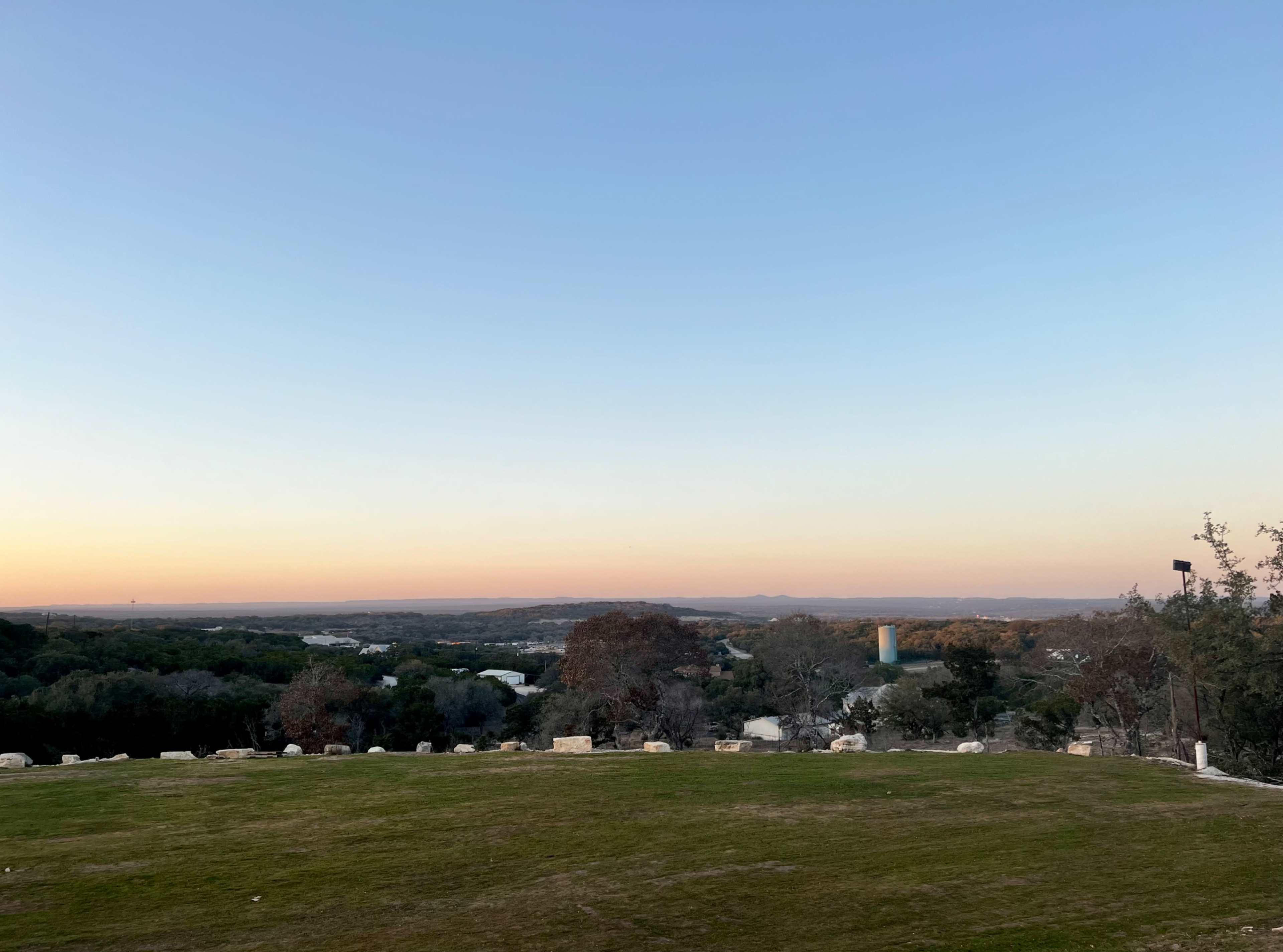 A wide view of a landscape at dusk, featuring rolling hills, trees, and a water tower in the distance under a clear sky.