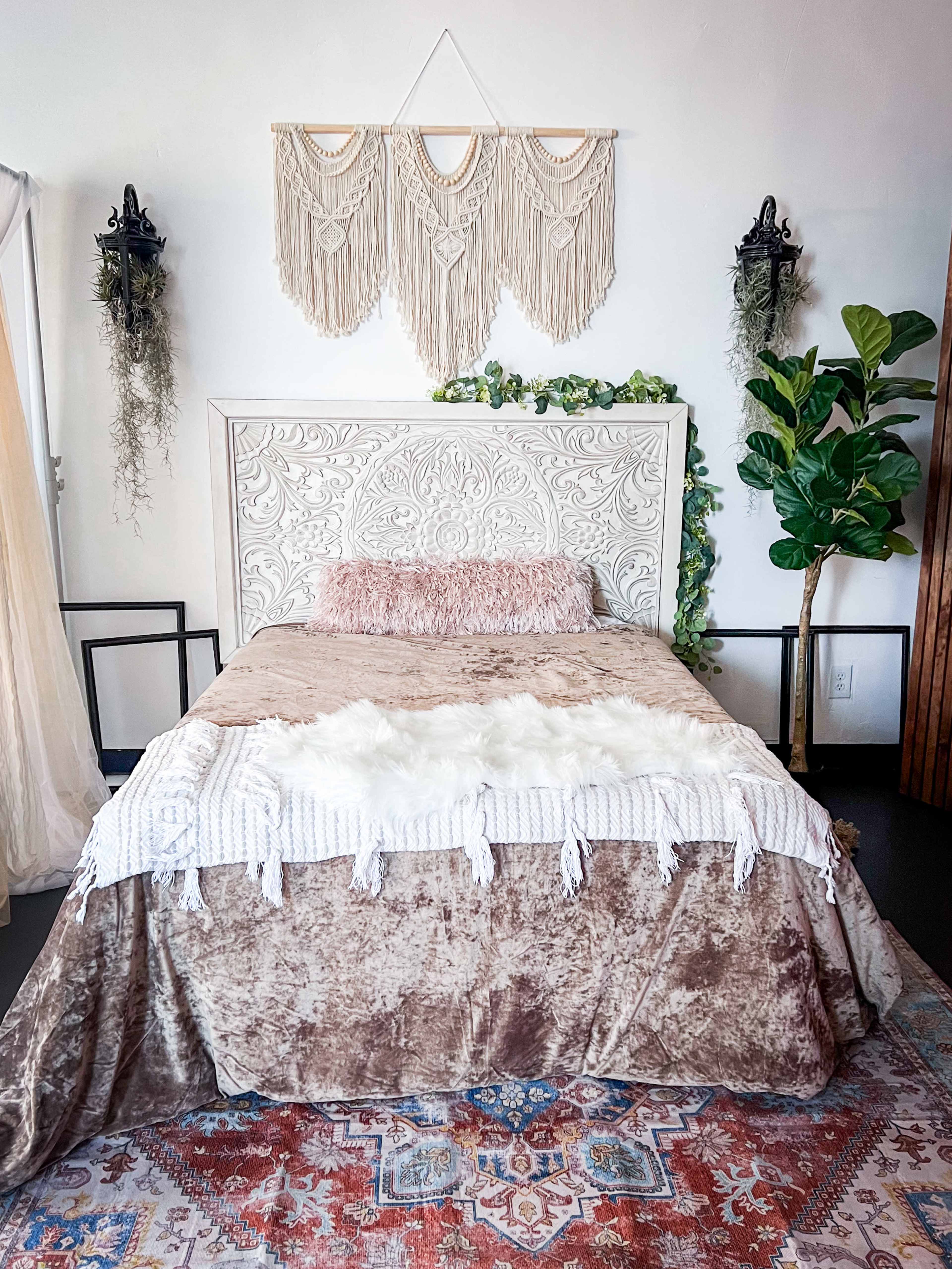 A bedroom featuring a bed with a decorative headboard, layered bedding, wall hangings, and plants, set against a textured rug.