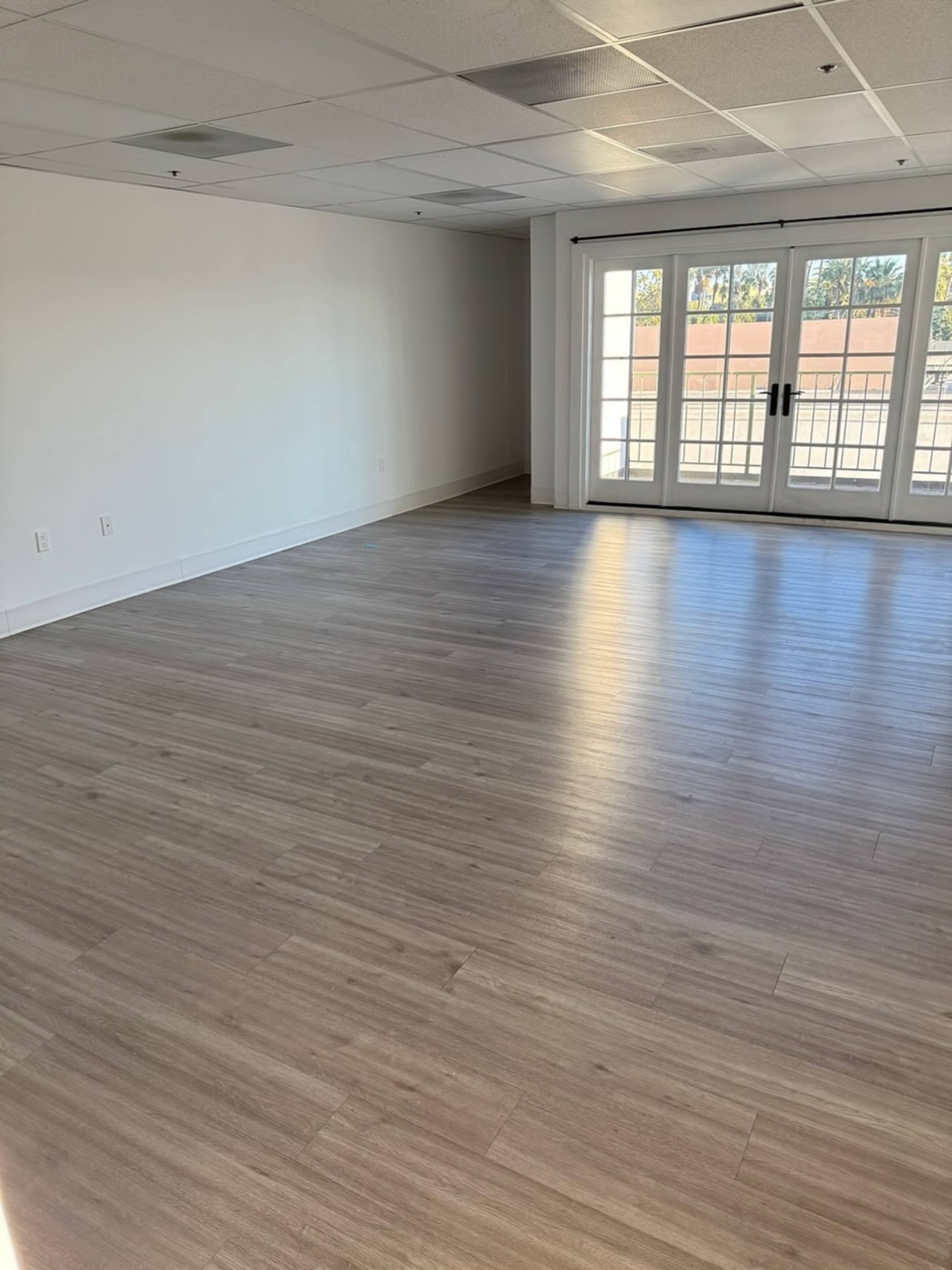 The image shows a spacious, empty room with wooden flooring and large windows that open to a balcony.