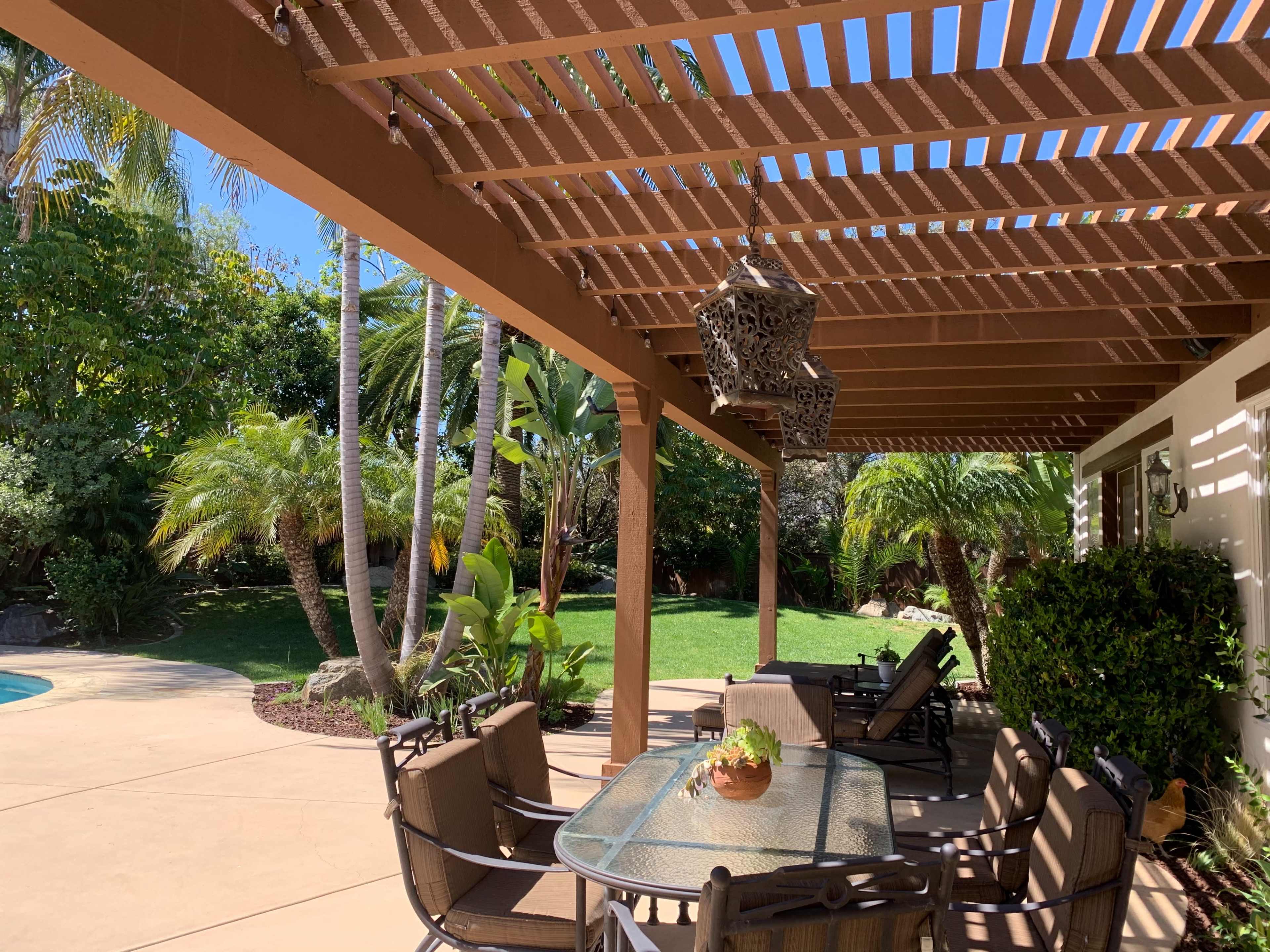 A patio area features a glass table surrounded by chairs, with a pergola overhead and tropical plants in the background.
