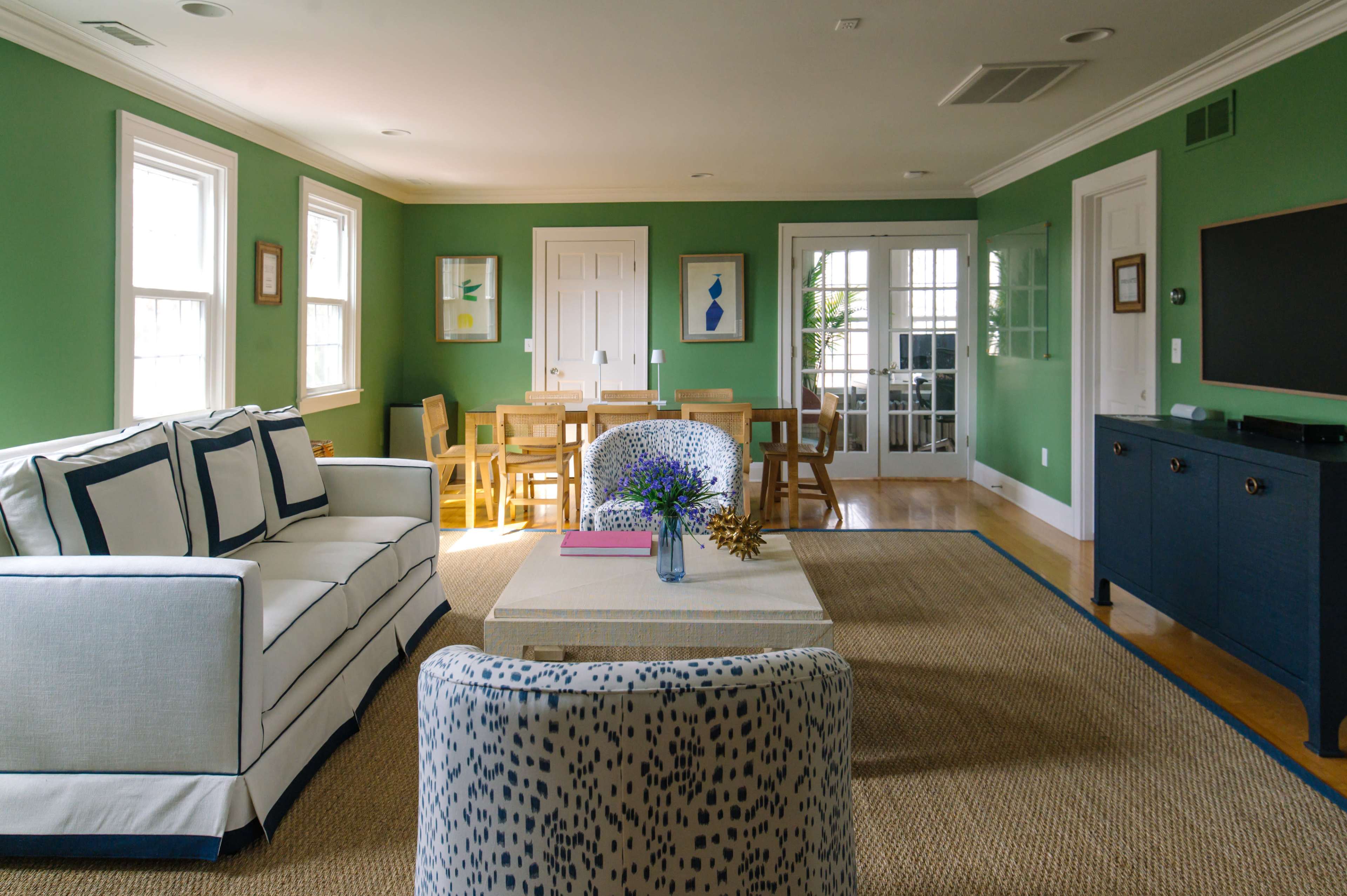 A living room with green walls, a white couch, a coffee table, a dining area with wooden chairs, and a television against a dark blue cabinet.