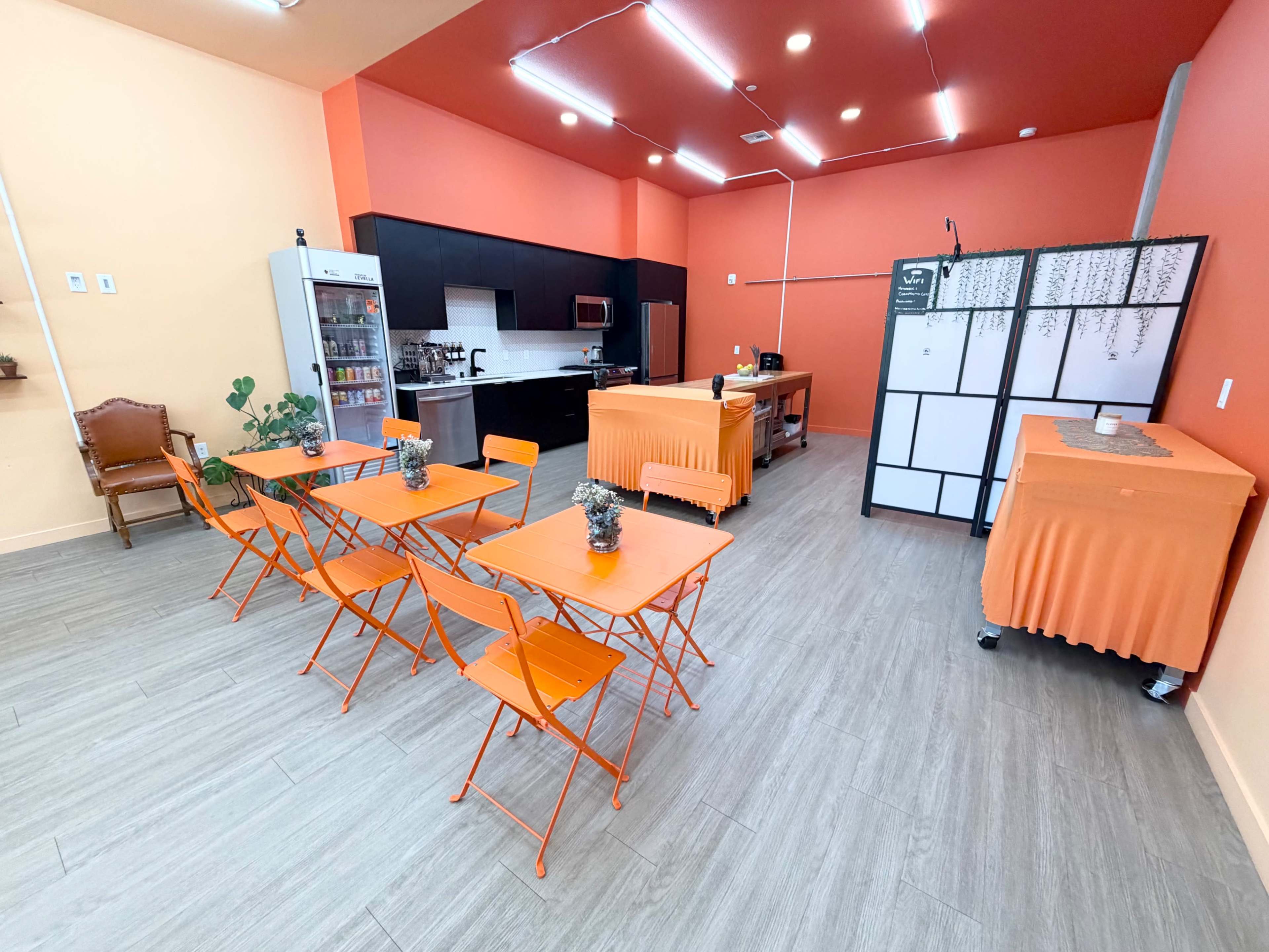 The image shows a brightly colored room with orange walls featuring a kitchen area, several orange metal tables and chairs, and a small counter space with a fridge and equipment.