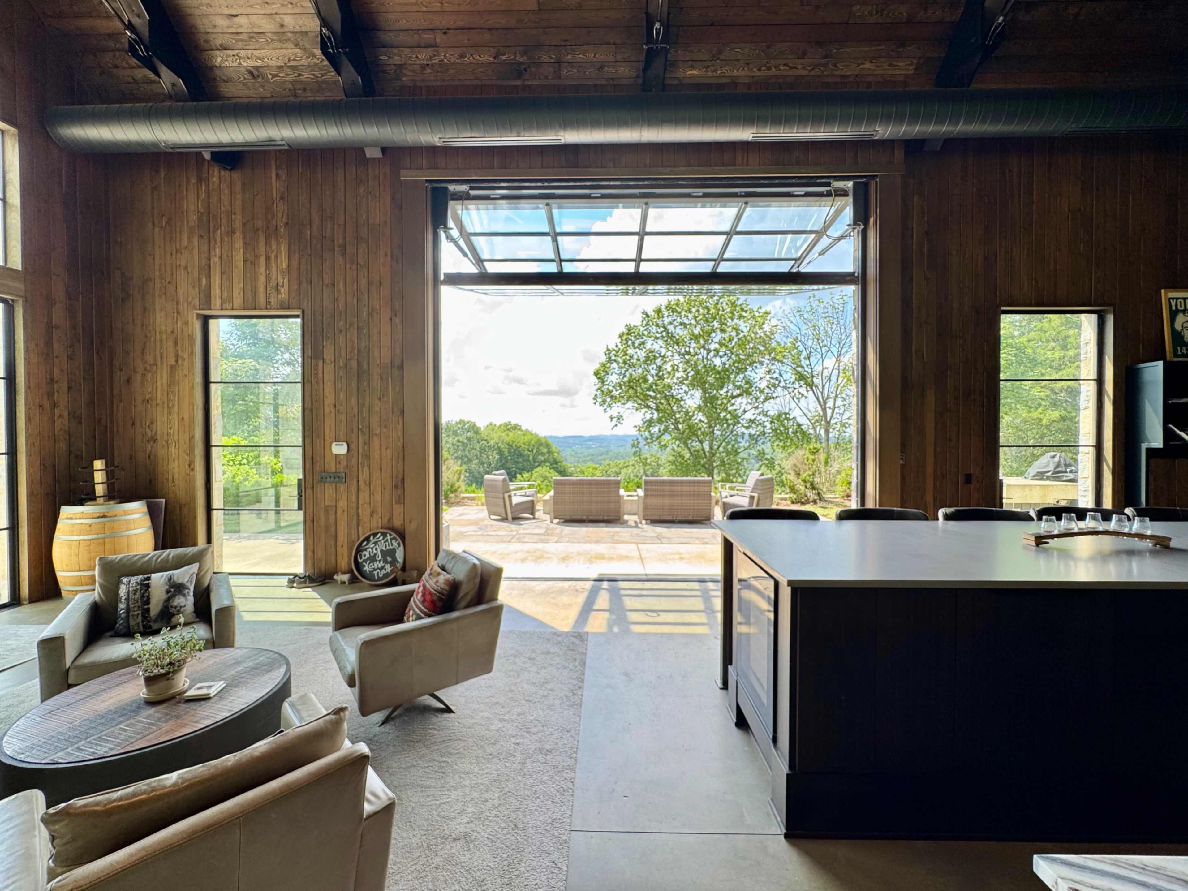 A modern interior space features a large open area with wood-paneled walls, a kitchen island, and a panoramic view of an outdoor patio and lush landscape through expansive glass doors.