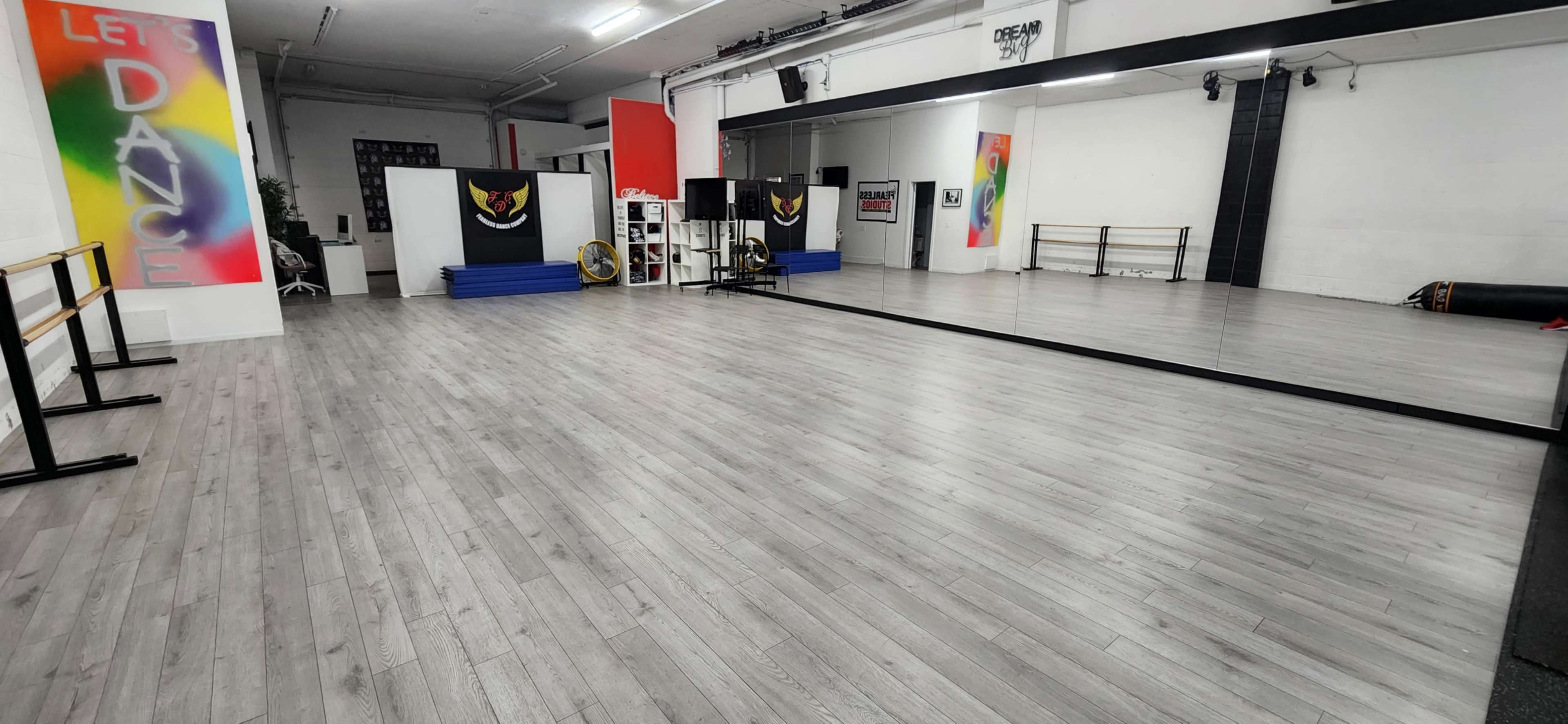 The image shows a spacious dance studio with gray wooden flooring, mirrors, and various exercise equipment arranged in a well-lit environment.
