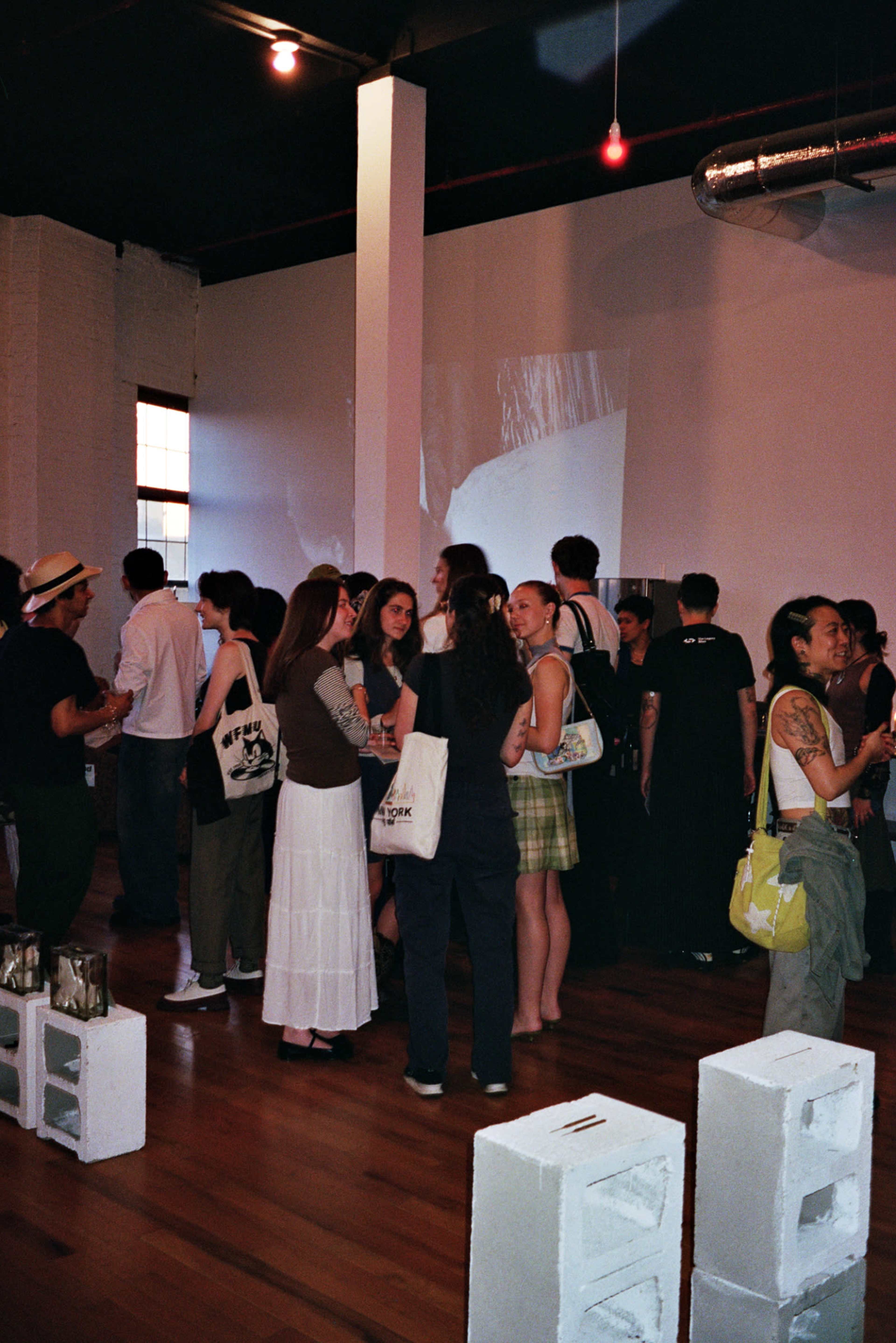 A group of people mingles at an art gallery event, surrounded by white walls and wooden floors, with video projections in the background.