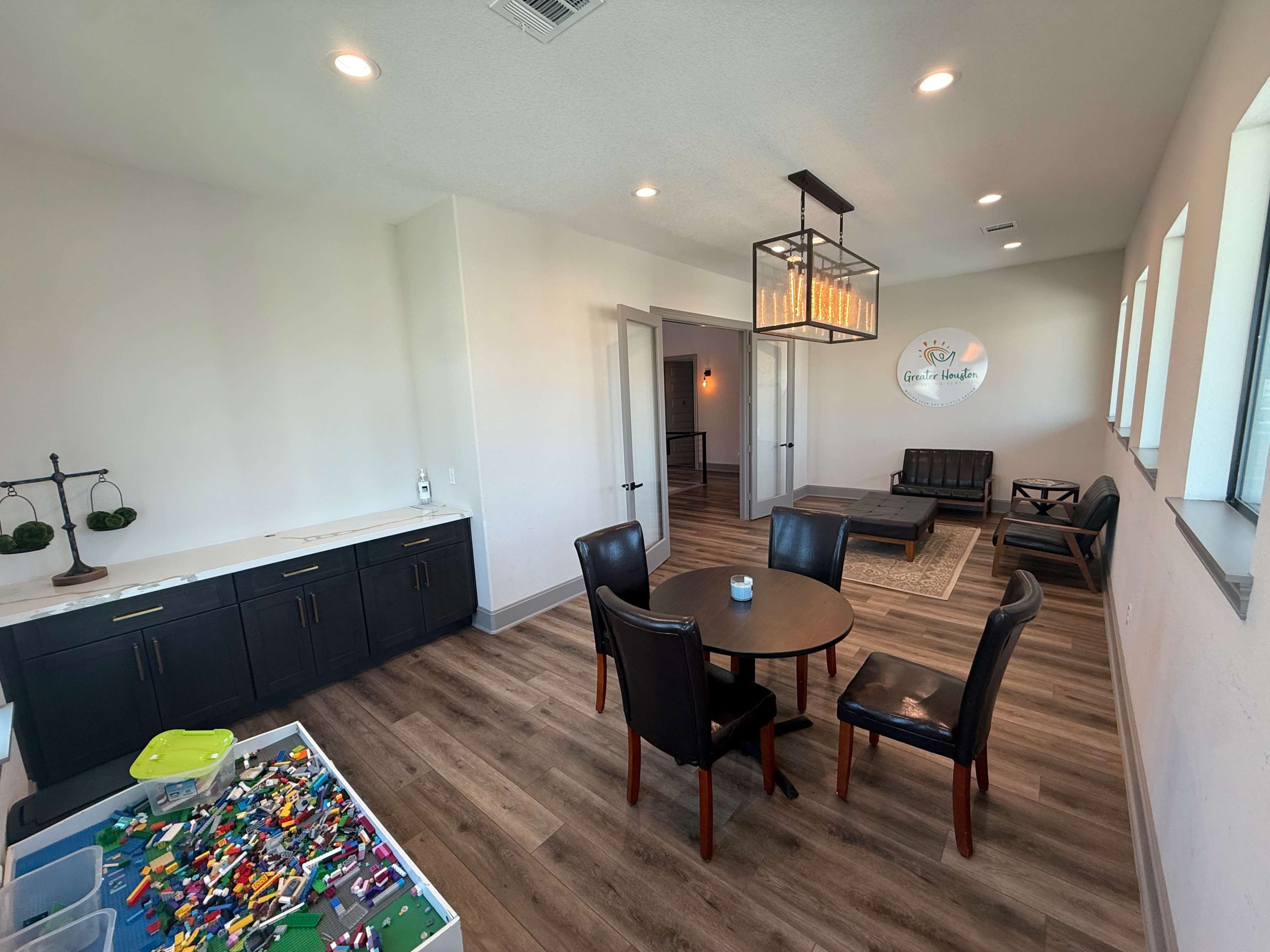 The image shows a bright, modern room with a small table and chairs, a black cabinet, and a play area filled with colorful building blocks.