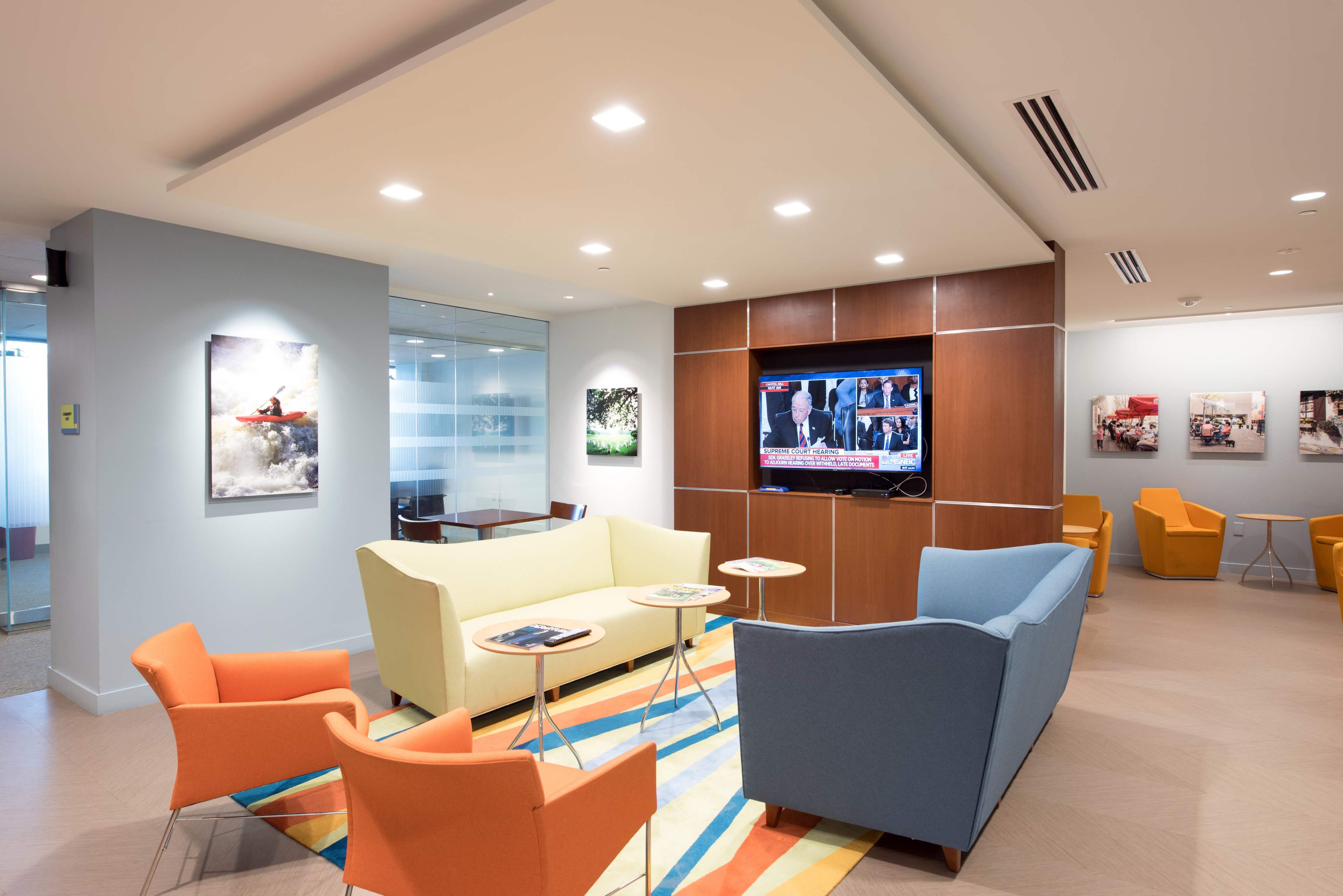 A modern waiting area features two sofas and several colorful chairs arranged around small tables, with a television displaying news in the background.