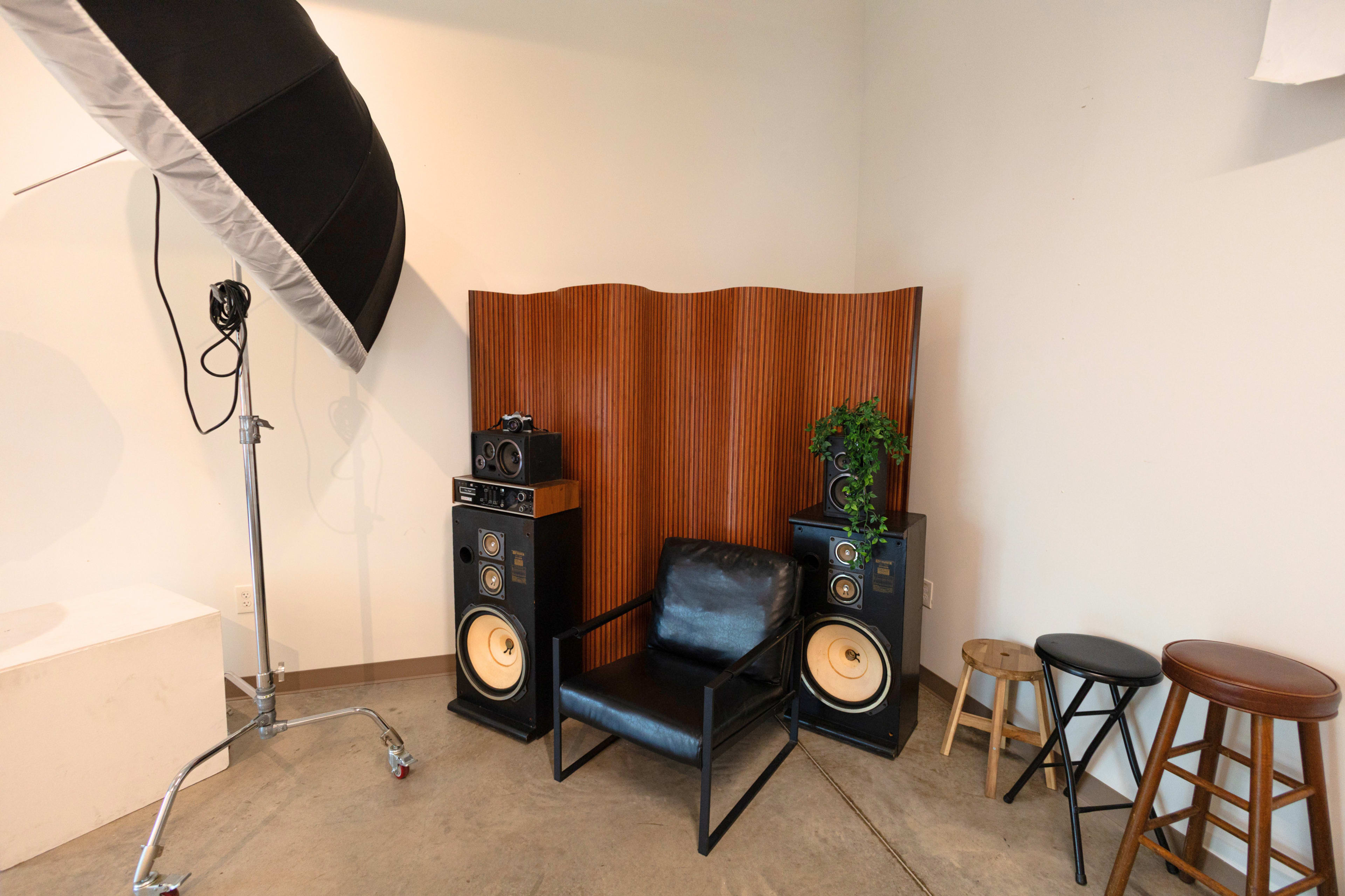 The image shows a corner of a room with a black chair in front of a wooden acoustic panel, flanked by vintage speakers and a plant, with a large photo umbrella reflector on a stand nearby.