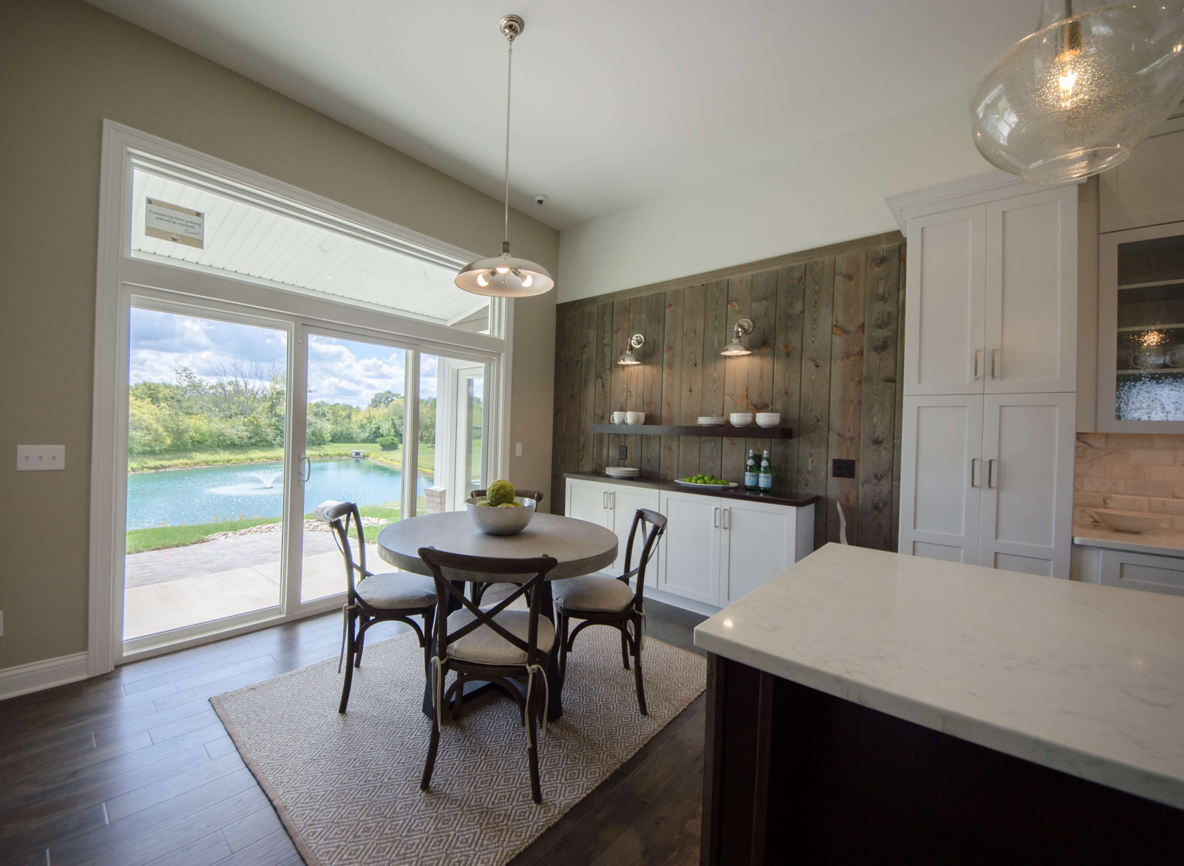 A round dining table with four chairs is positioned near a large window that overlooks a scenic outdoor view by the water.