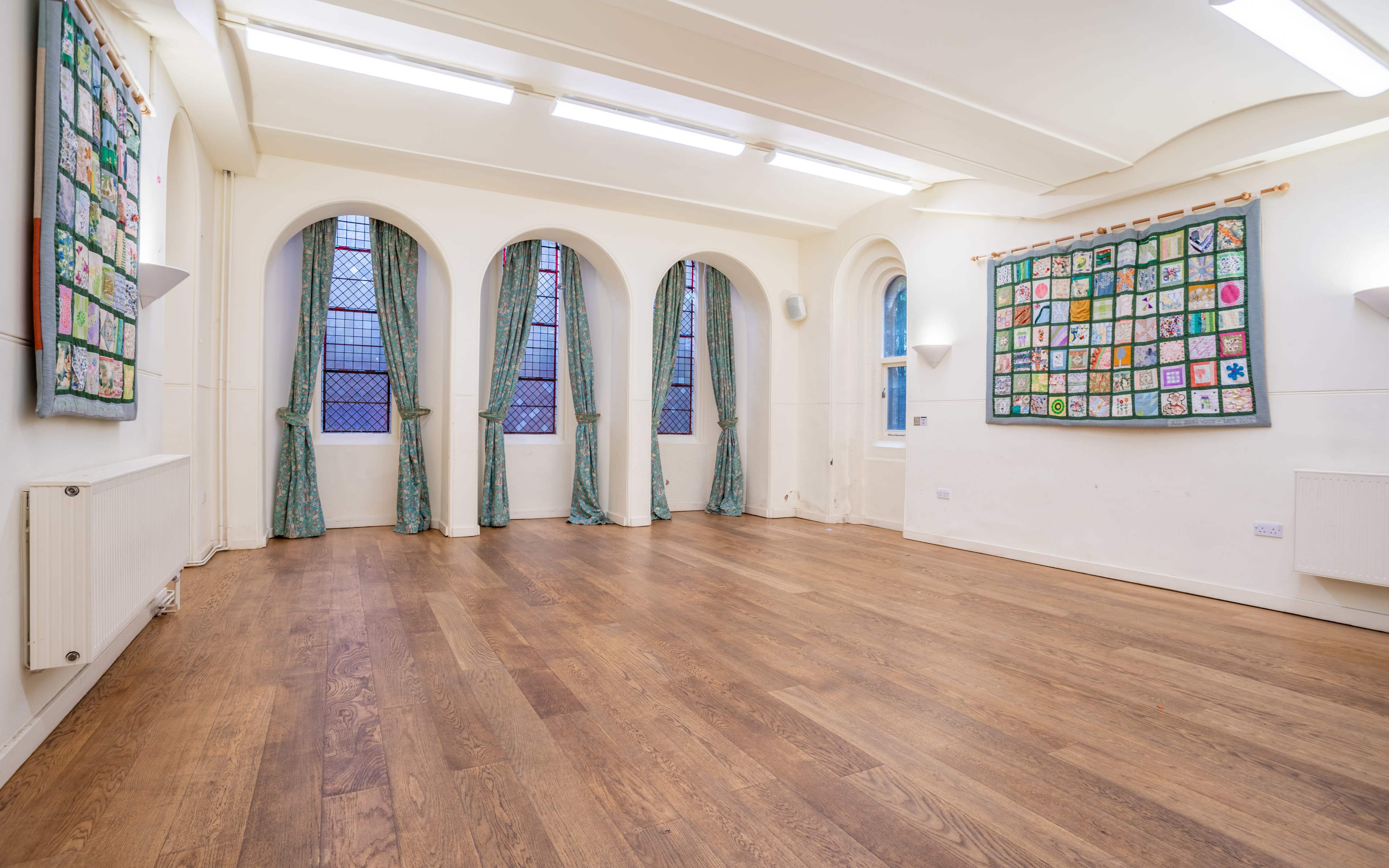 A spacious, well-lit room with high ceilings, large arched windows, and colorful textile artworks hanging on the walls.