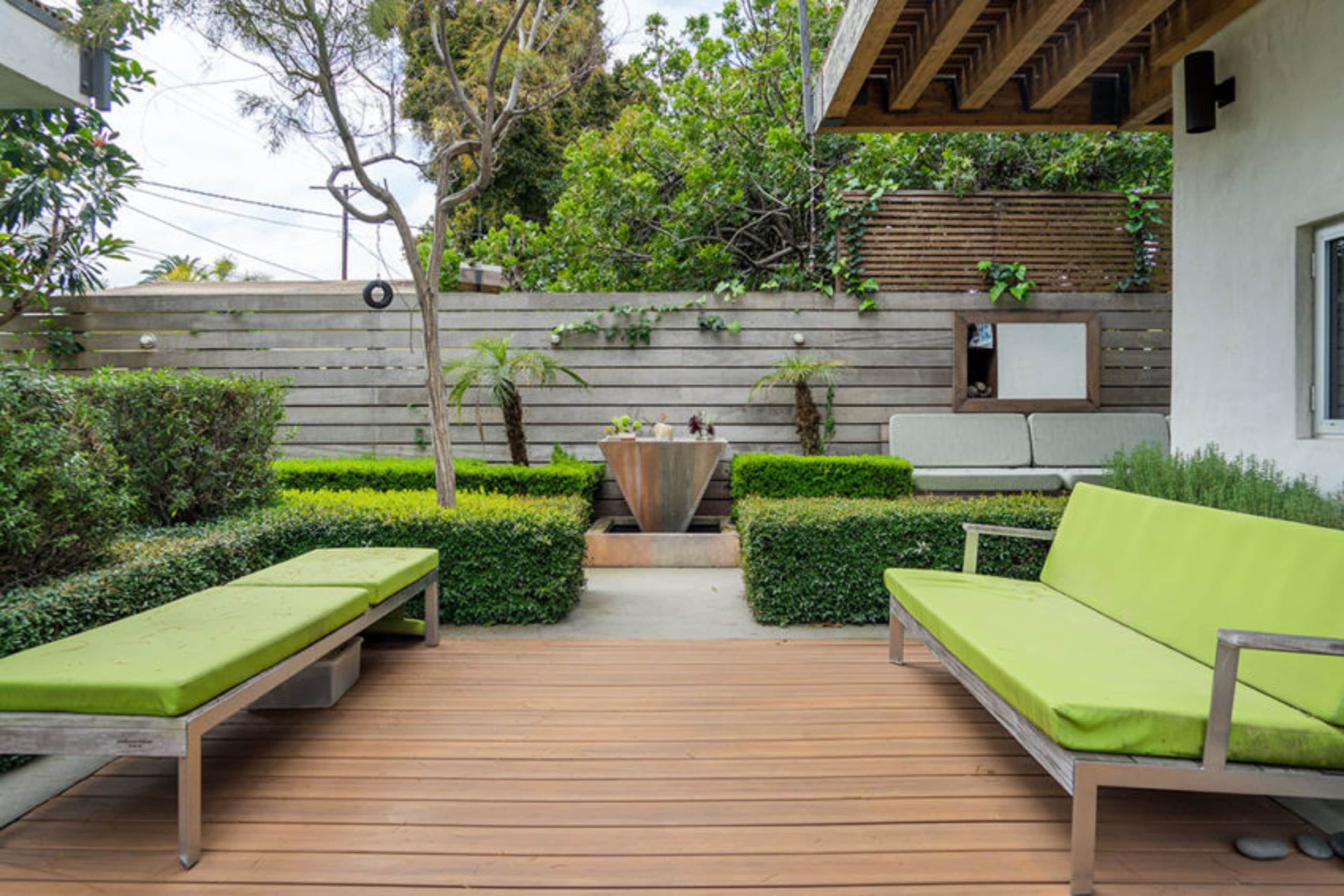 A modern outdoor patio features two green benches, a central water feature, and landscaped greenery surrounded by wooden fencing.