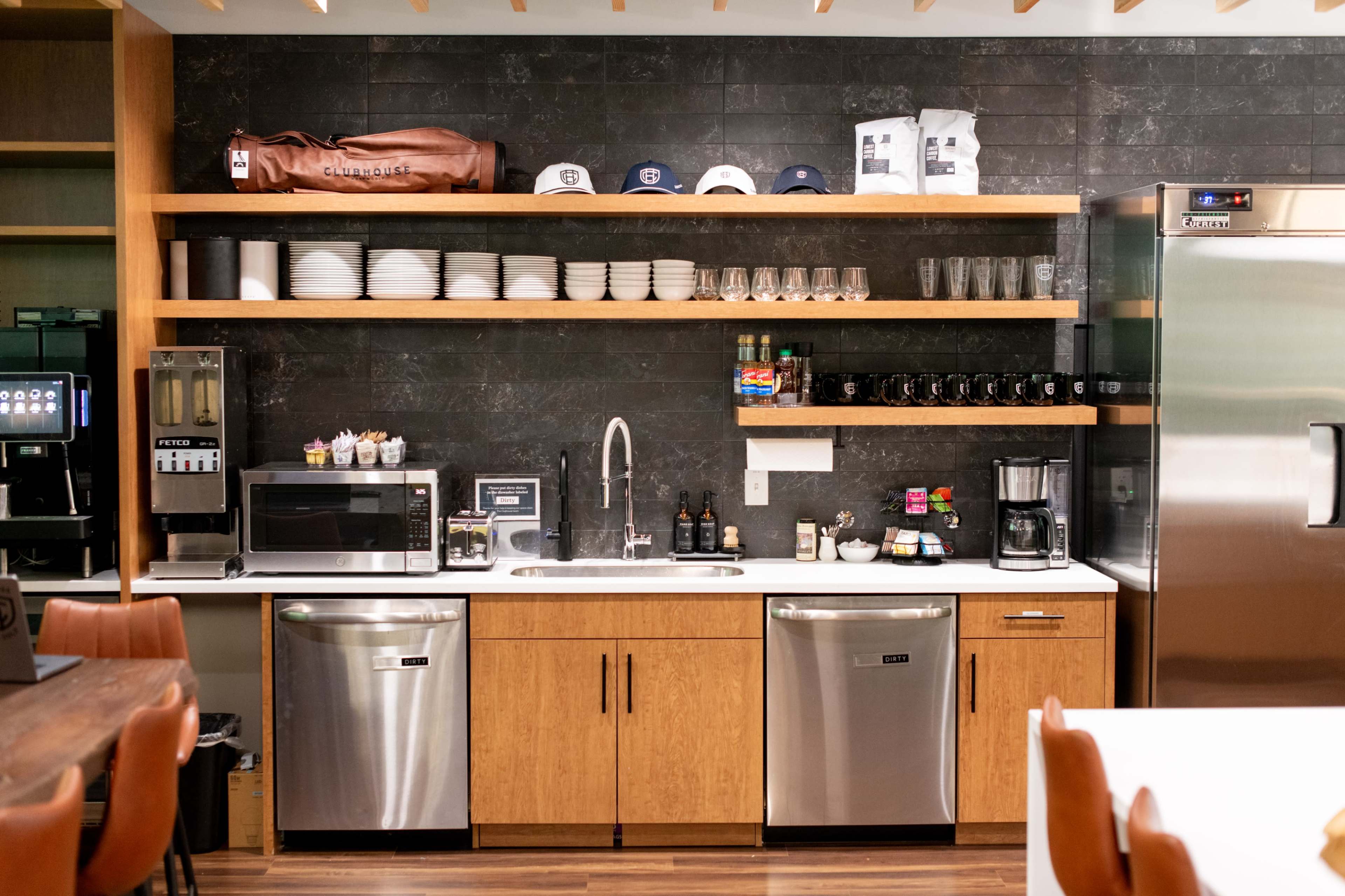 The image shows a modern kitchen area featuring a microwave, sink, coffee machine, and stainless steel appliances, with shelves holding cups, glasses, and other kitchen items.