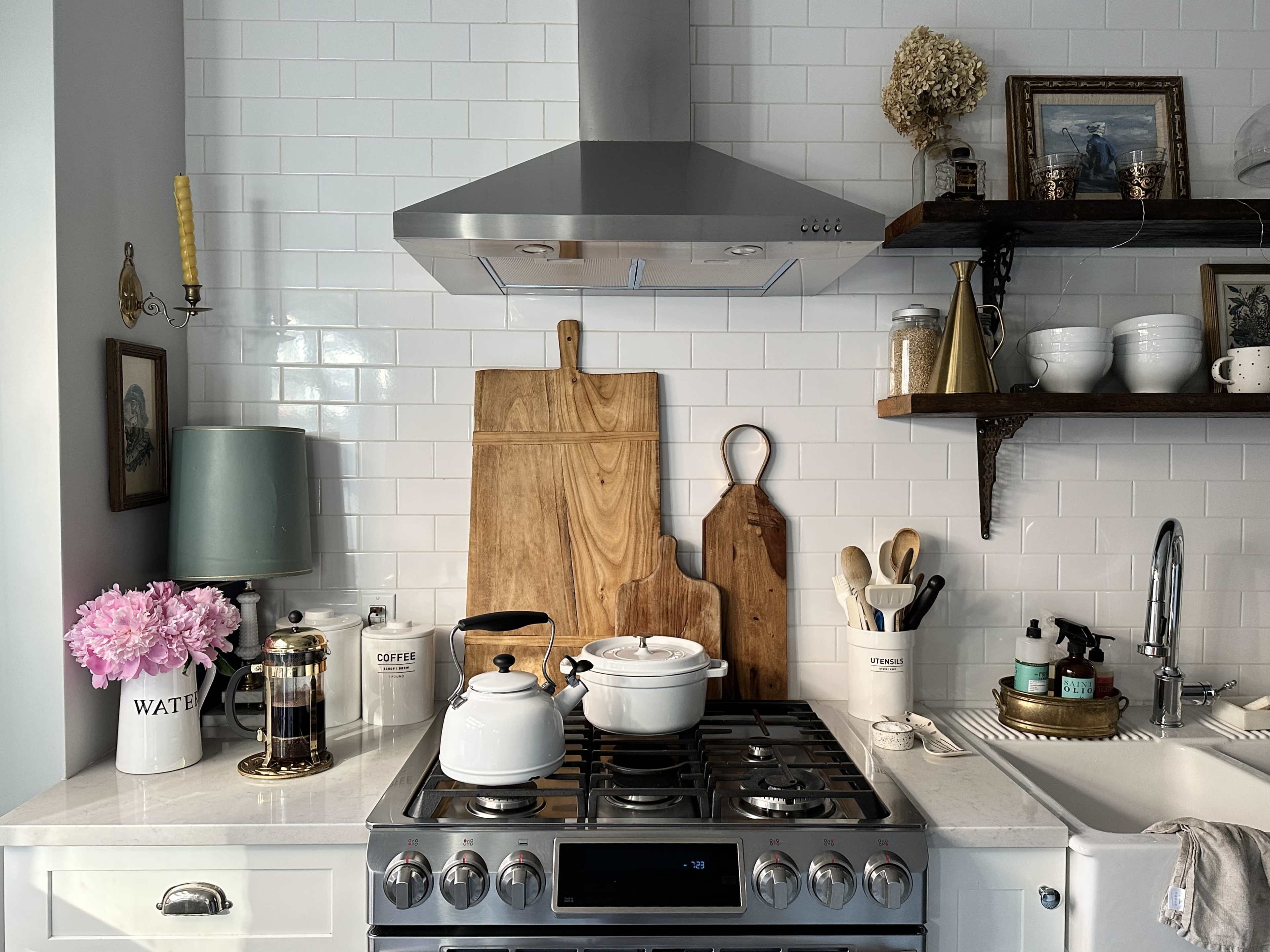 A modern kitchen features a stainless steel hood, wooden cutting boards, white cookware, and decorative accents, all arranged neatly on a marble counter.