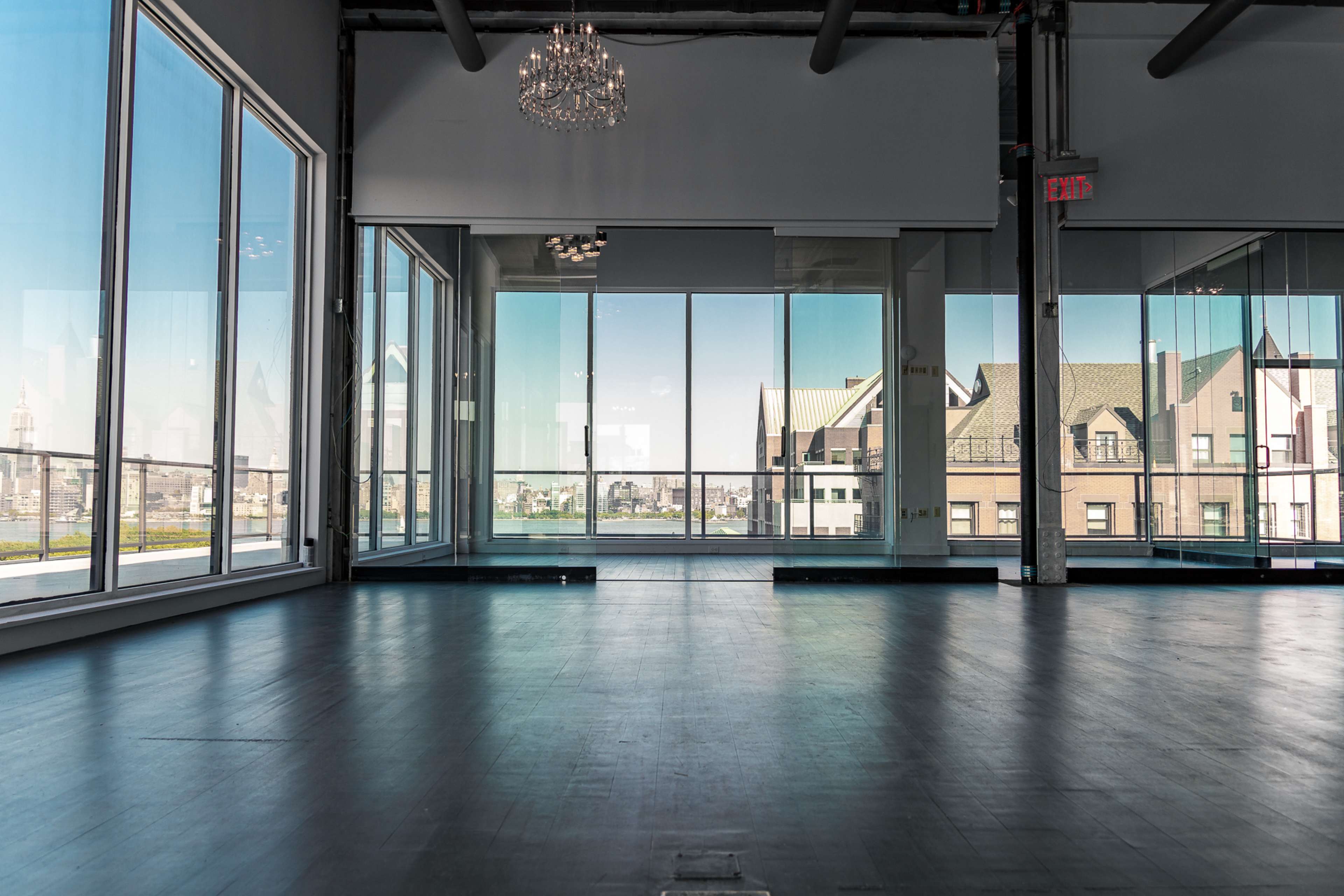 A spacious interior room features large windows overlooking city buildings and a chandelier above the empty wooden floor.
