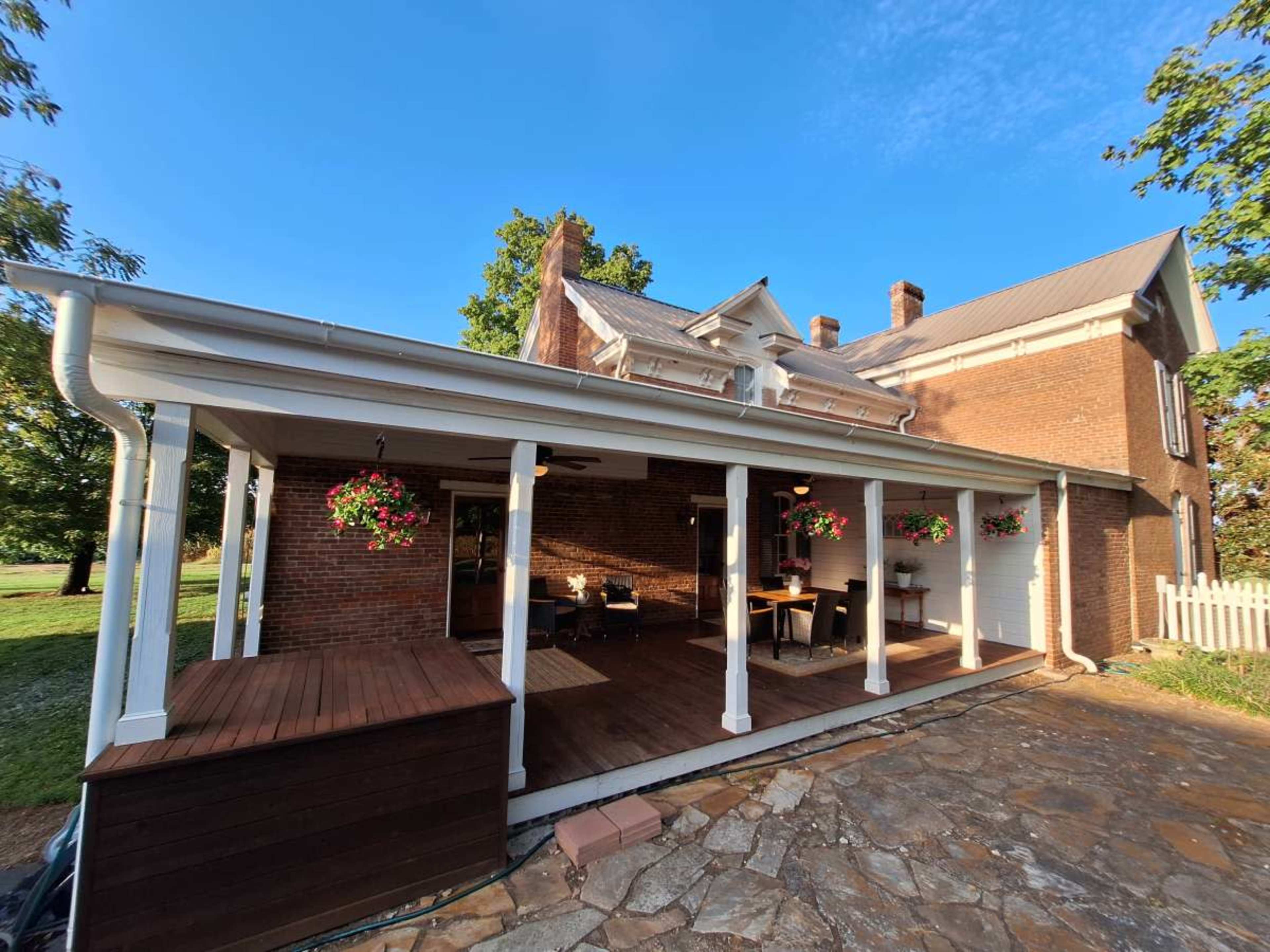 The image shows a brick house with a covered porch featuring wooden decking and hanging flower pots.
