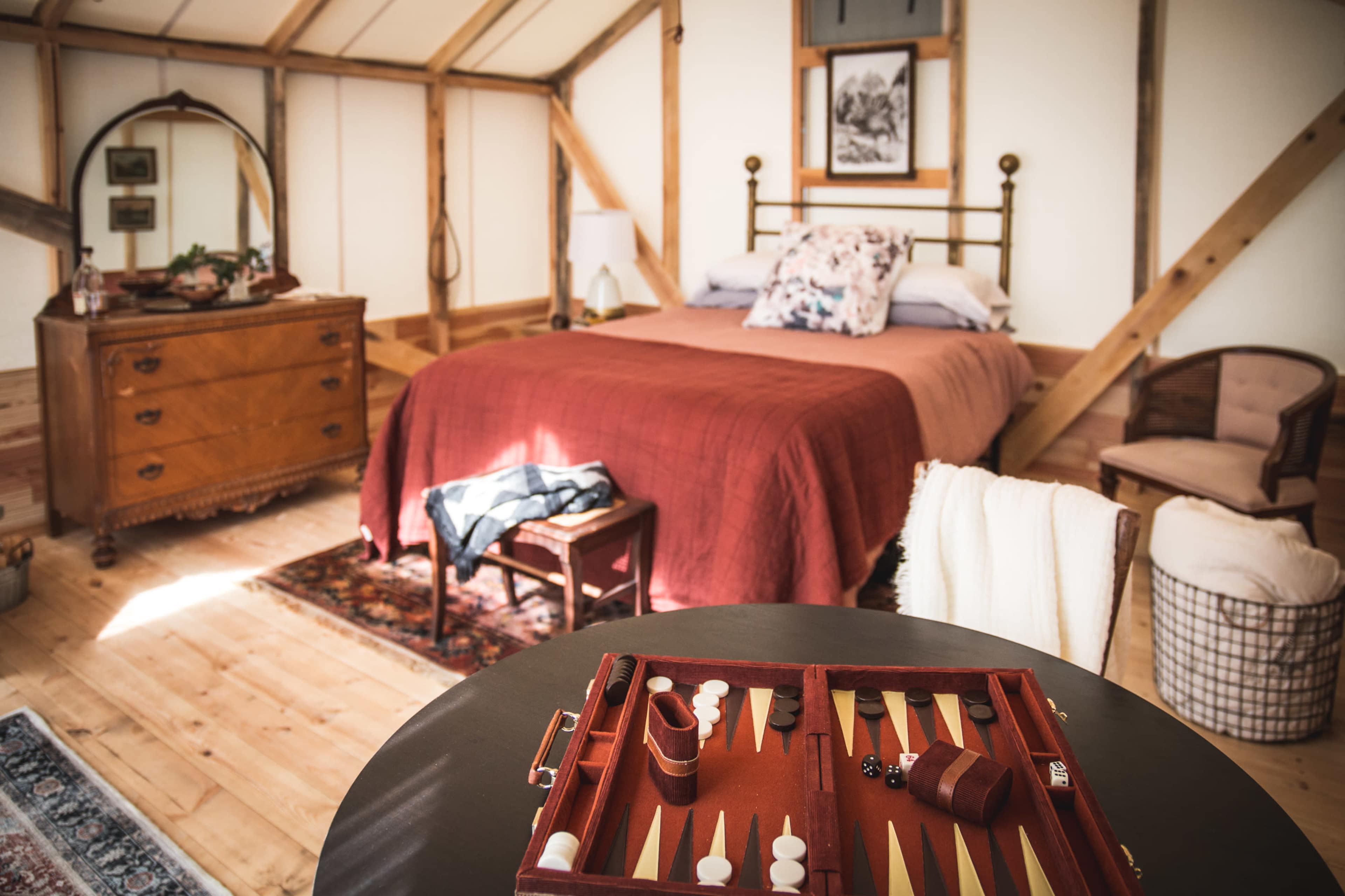 A cozy bedroom with a bed dressed in a burgundy bedspread, a vintage dresser, and a backgammon set placed on a round table.