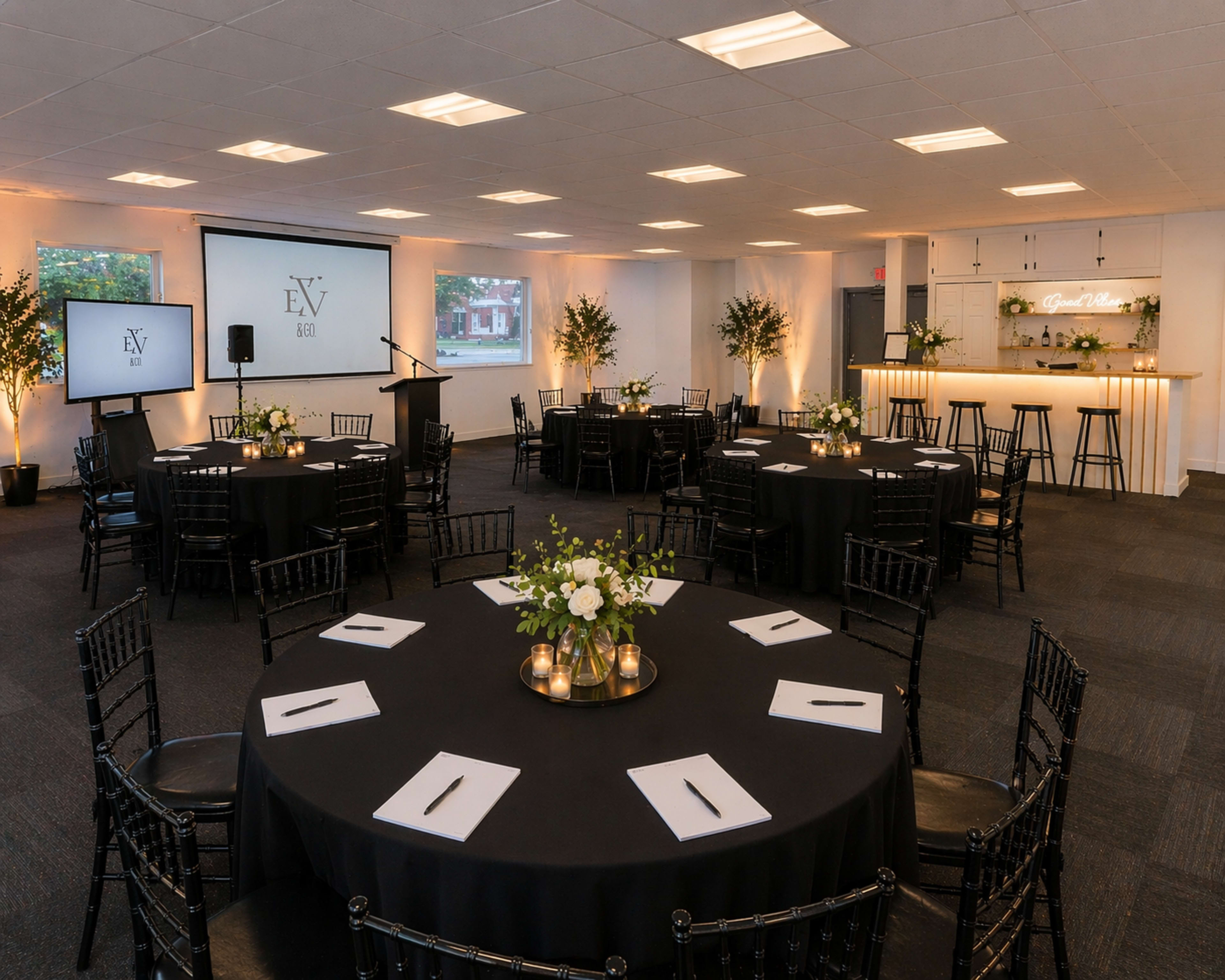 The image shows a spacious event room equipped with round tables covered in black cloth, each with decorative centerpieces, and a bar area illuminated with soft lighting.