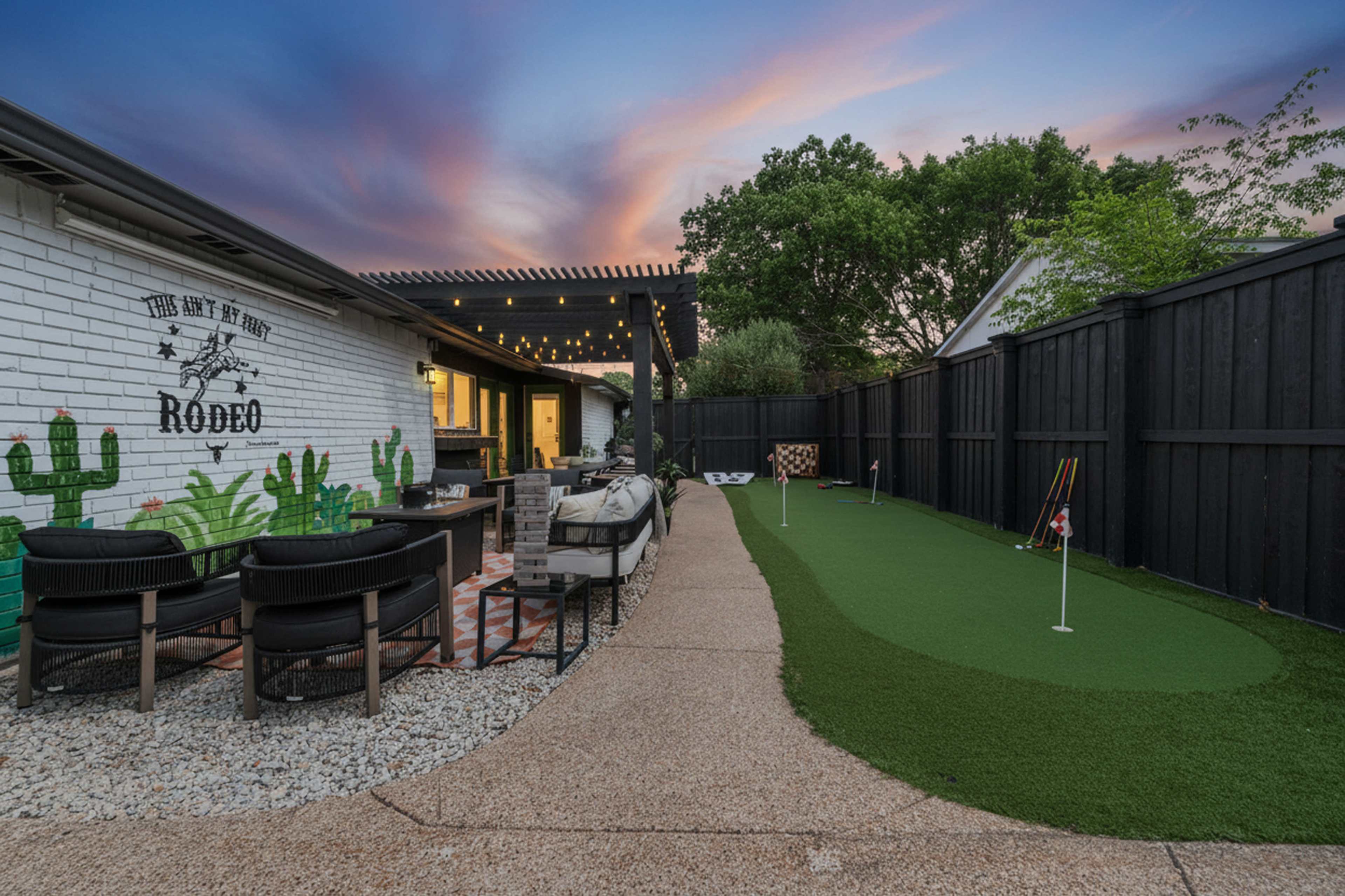 A modern backyard features a putting green alongside a patio area with lounge seating and a mural of cacti on a white brick wall.
