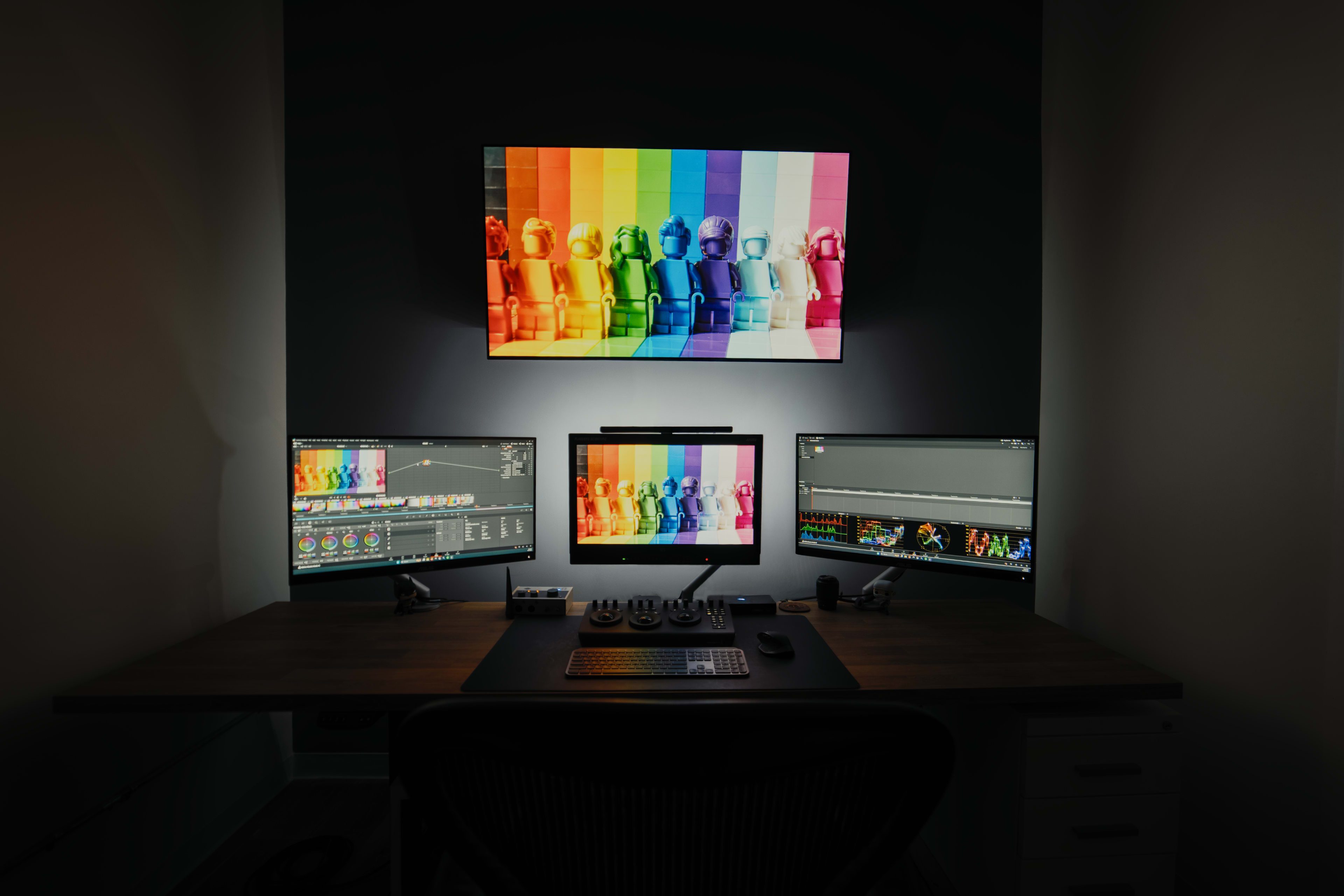 The image depicts a modern workspace with three computer monitors displaying colorful graphics, set against a wall with a large screen showing a similar vibrant design.