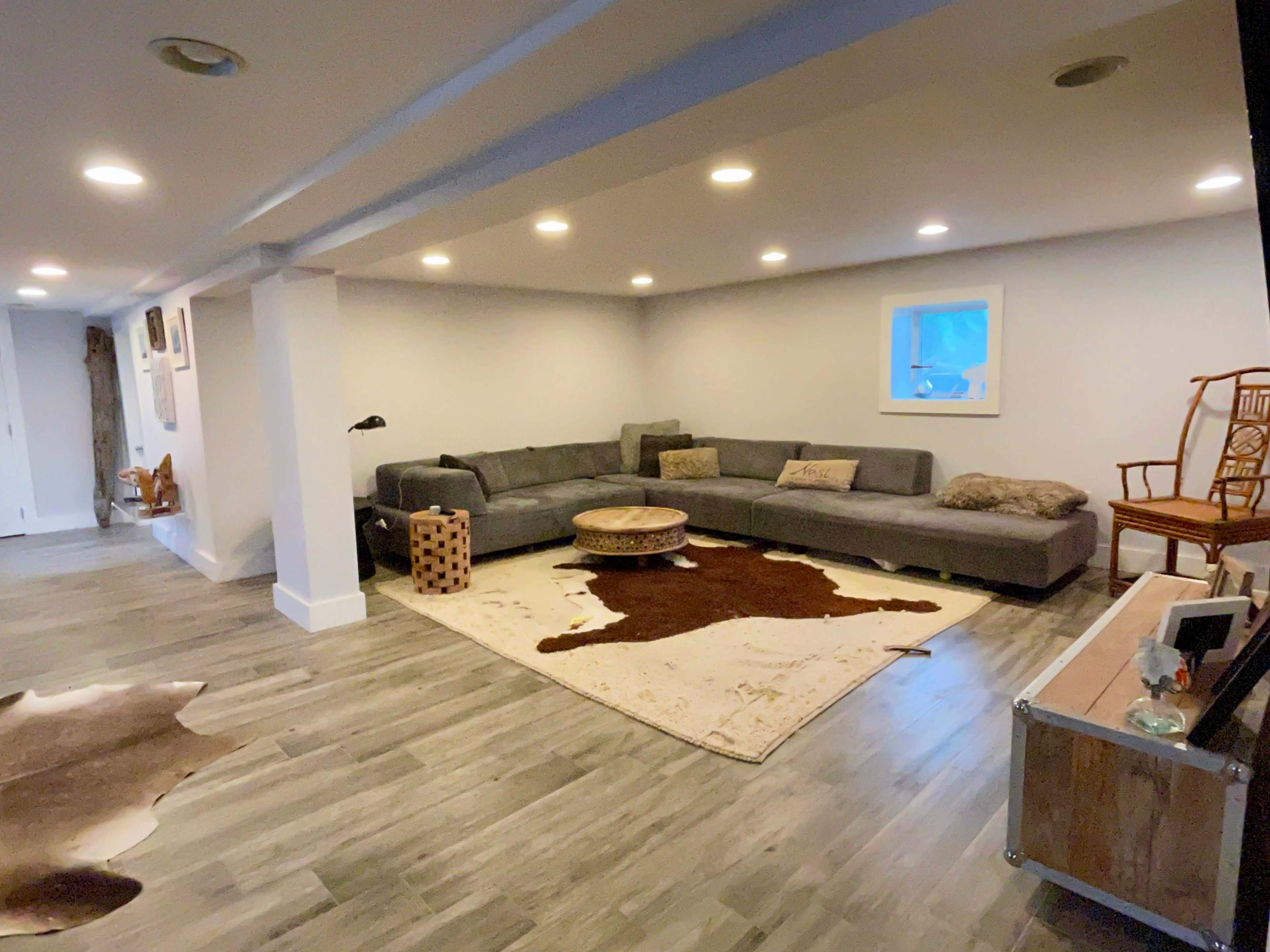 A spacious basement living area features a gray sectional sofa, a round coffee table, and a decorative area rug on a wooden floor, illuminated by recessed lighting.