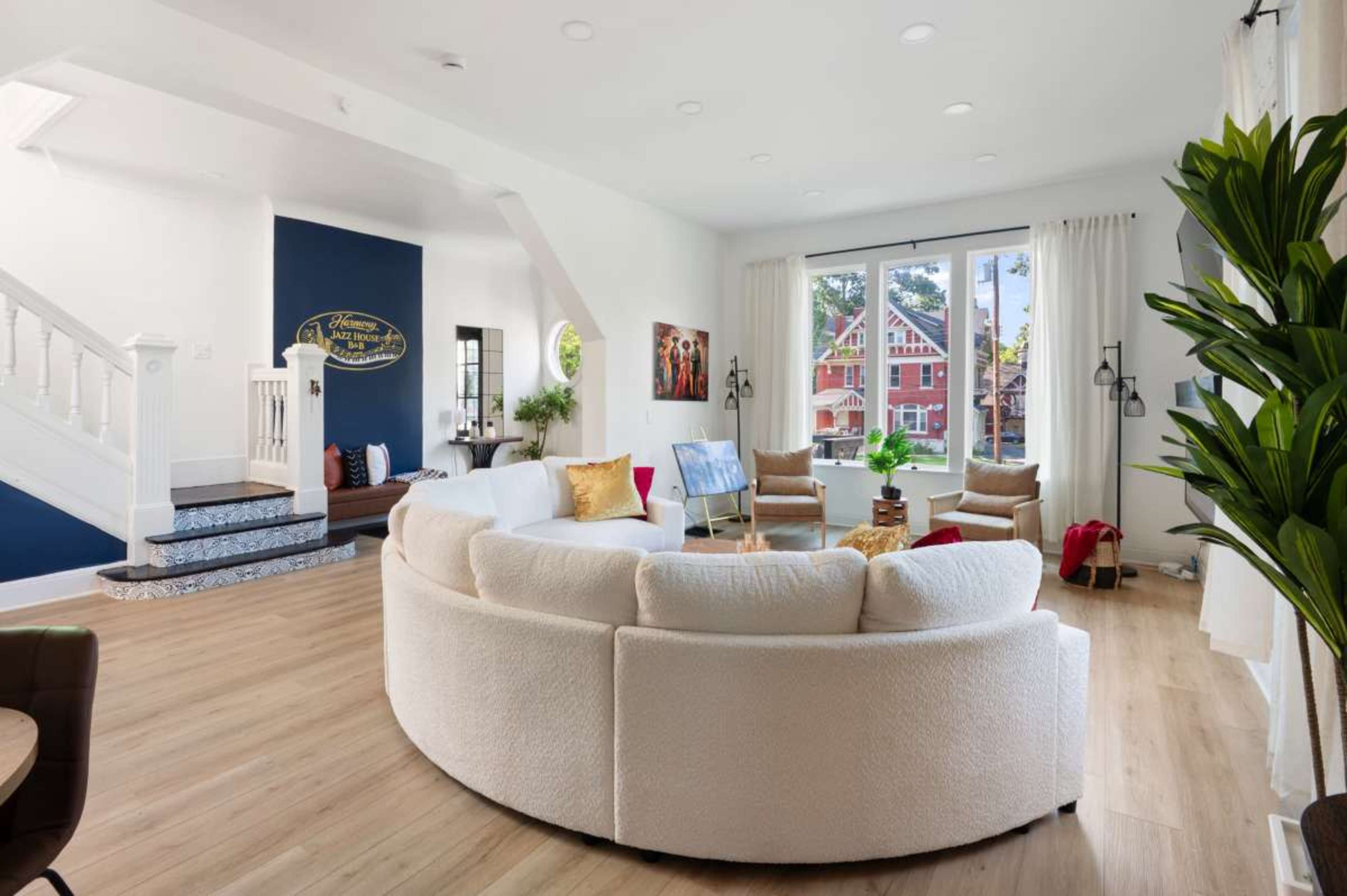 A circular, upholstered sofa is positioned in a bright living room, complemented by large windows that reveal a view of an exterior garden and staircase.