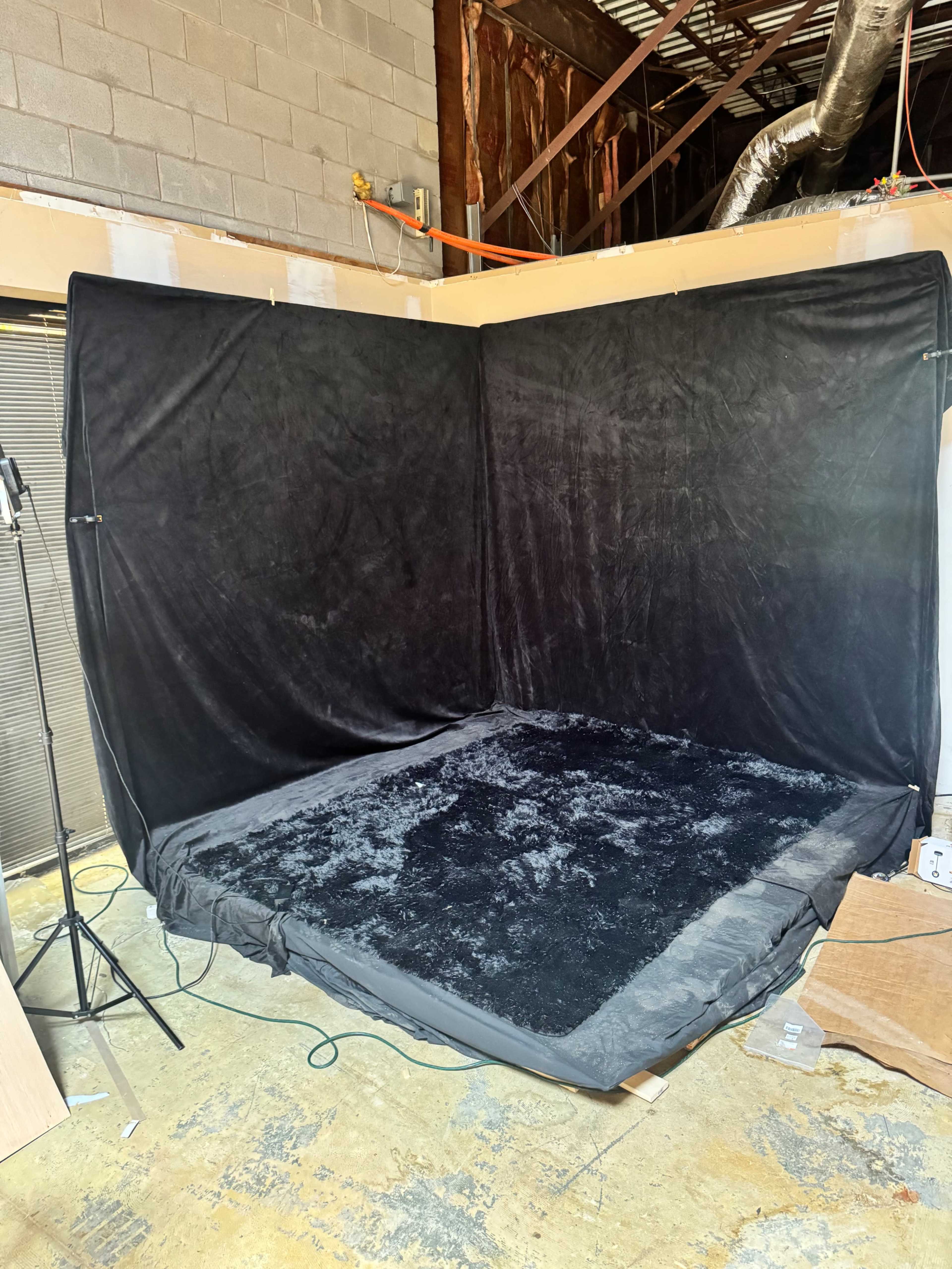 The image shows a corner of a room with a black fabric backdrop and a dark, carpeted area, set up for a photography or video session.