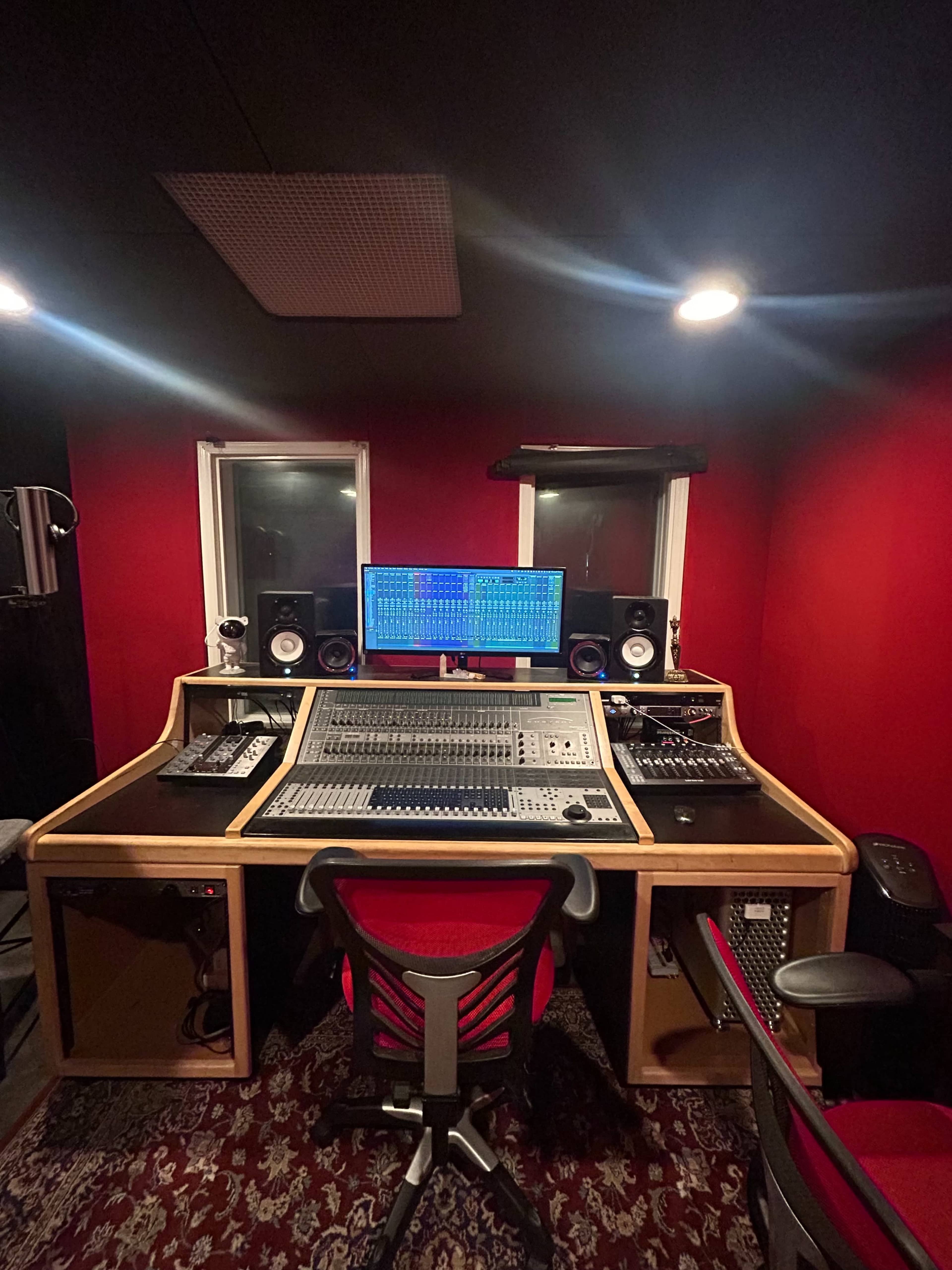 A recording studio features a large mixing console, computer monitor, and audio equipment set against red walls.