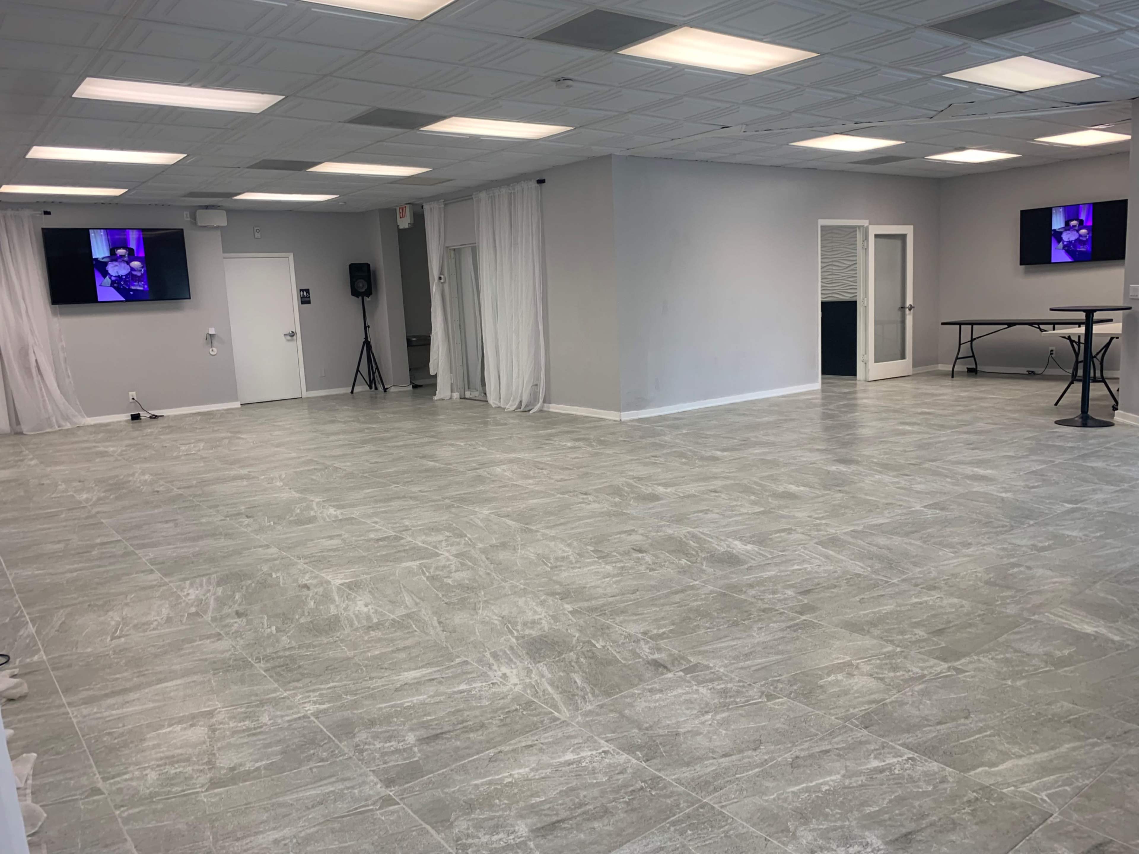 A spacious room with light gray tiled flooring, white walls, and two screens mounted on the walls, along with a folding table in the corner.