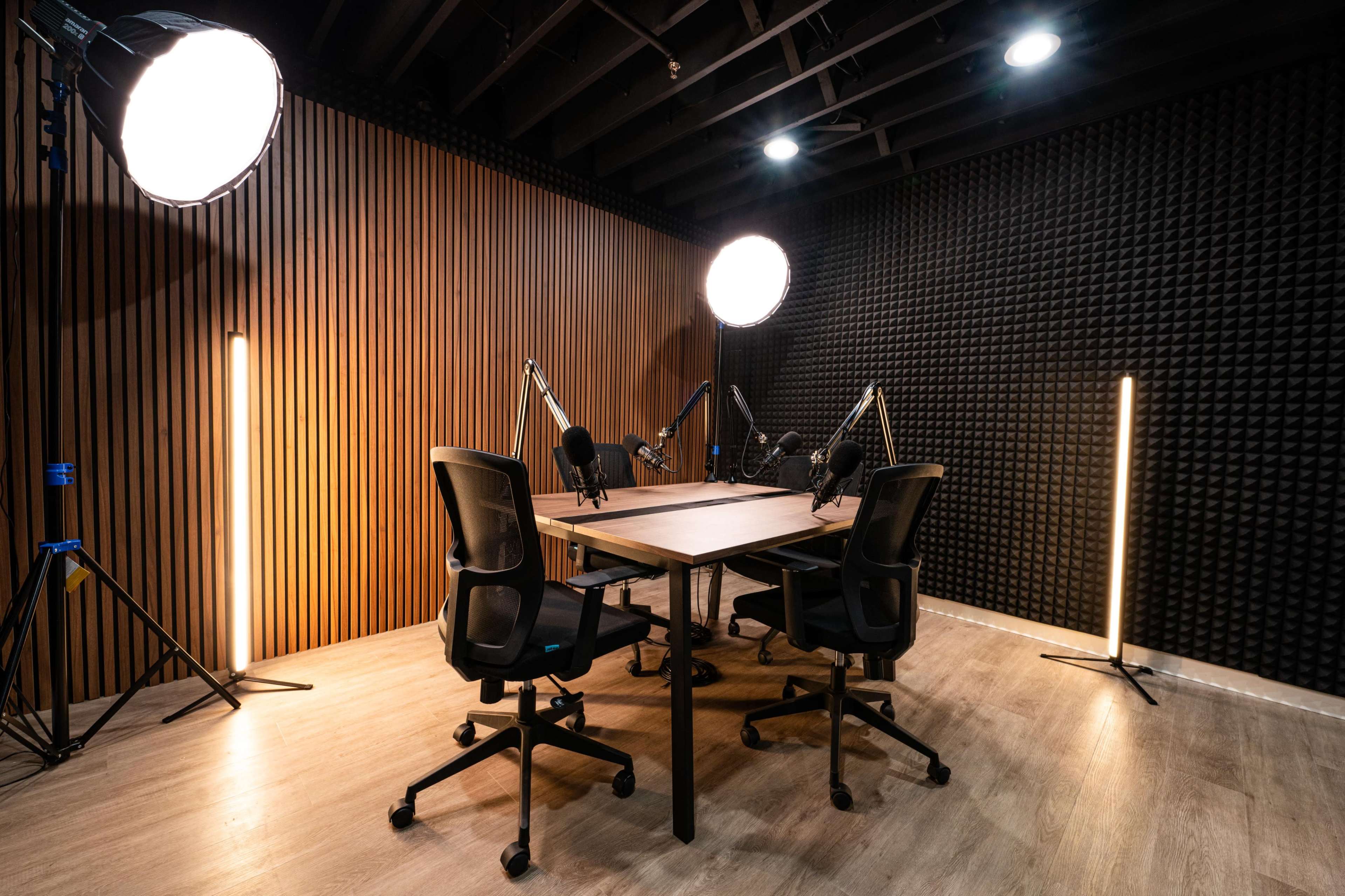 The image shows a recording studio with a wooden table, four chairs, and multiple professional microphones set up against a soundproofed wall.