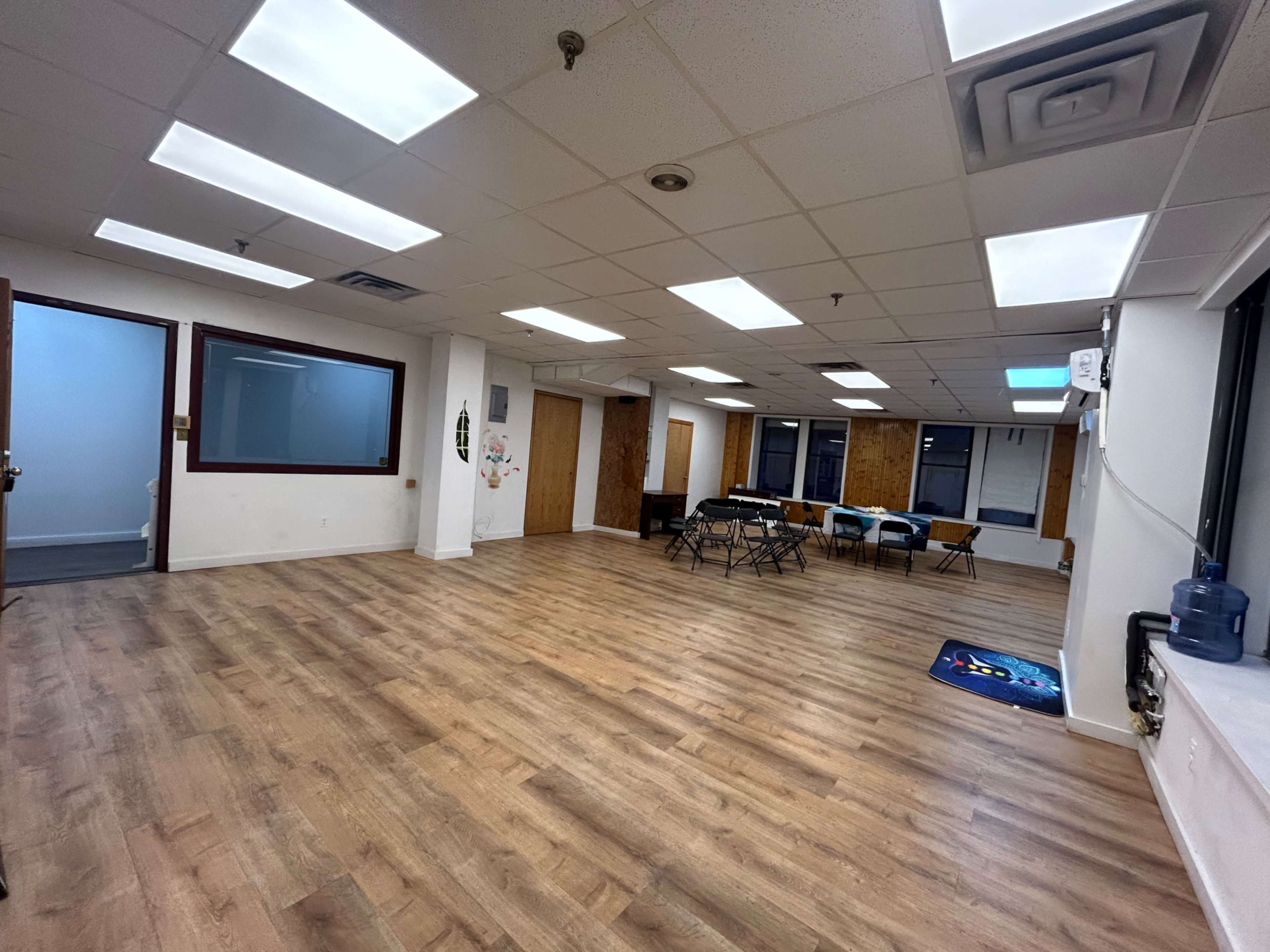 A spacious, bare room with wooden flooring, several chairs arranged around a table, and large windows providing natural light.