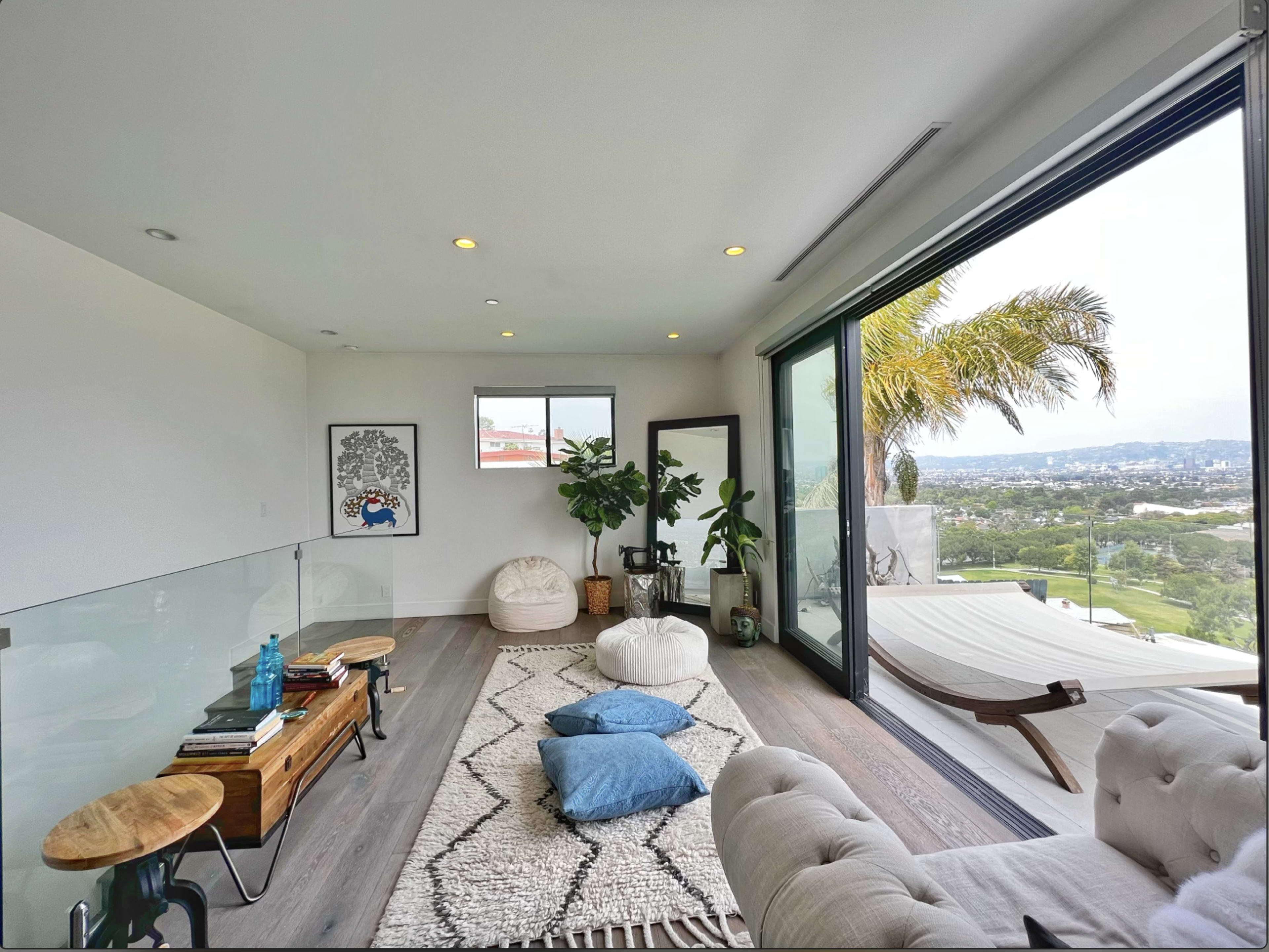 The room features a modern, minimalist design with large windows showcasing a view of the outdoors, and includes comfortable seating, a rug, and decorative plants.