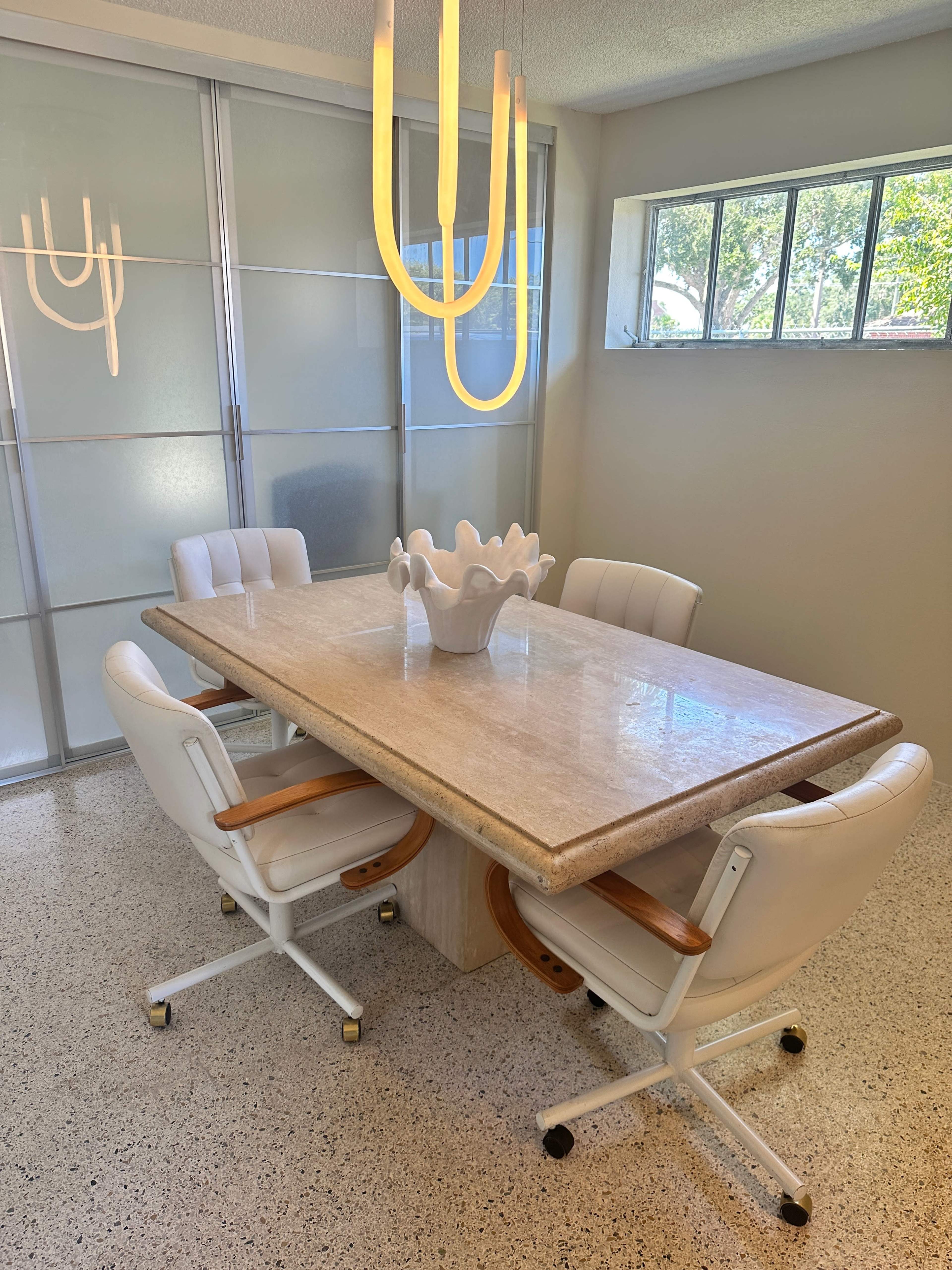 A modern dining area features a rectangular stone table surrounded by four white chairs and illuminated by a contemporary hanging light fixture.