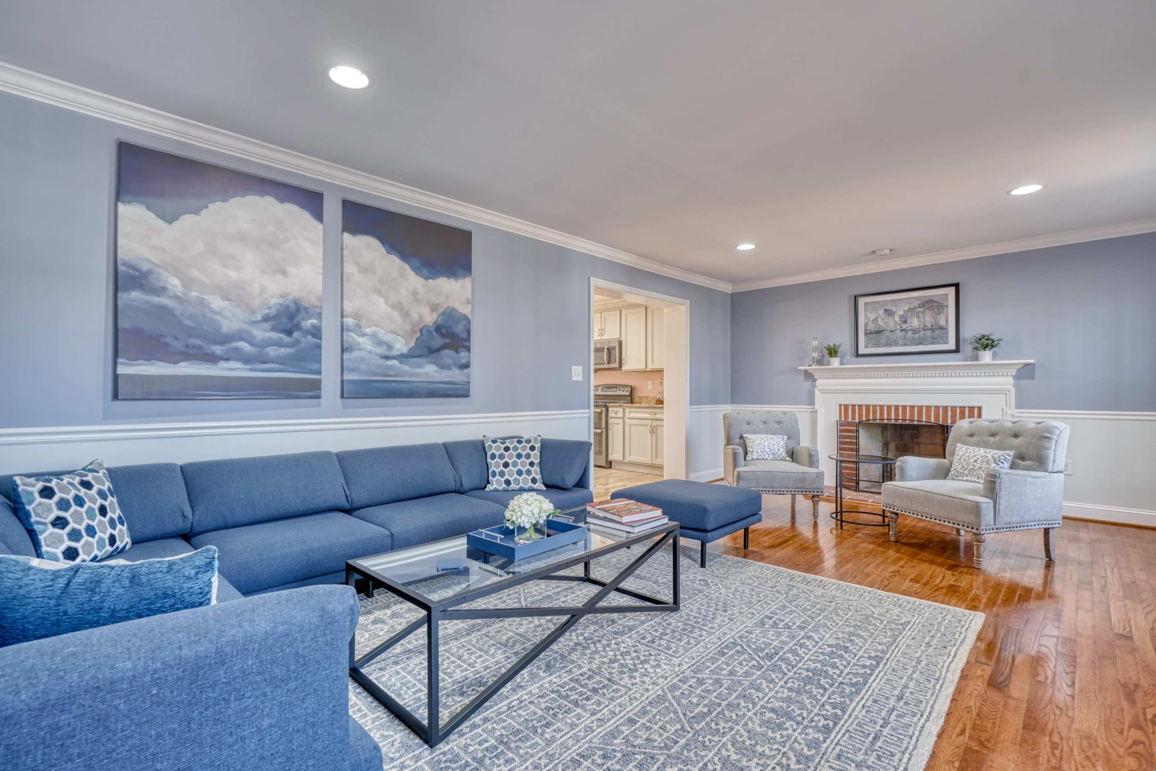 A living room with a blue sectional sofa, a glass coffee table, two armchairs, and a fireplace, all accented by wall art and a decorative rug.