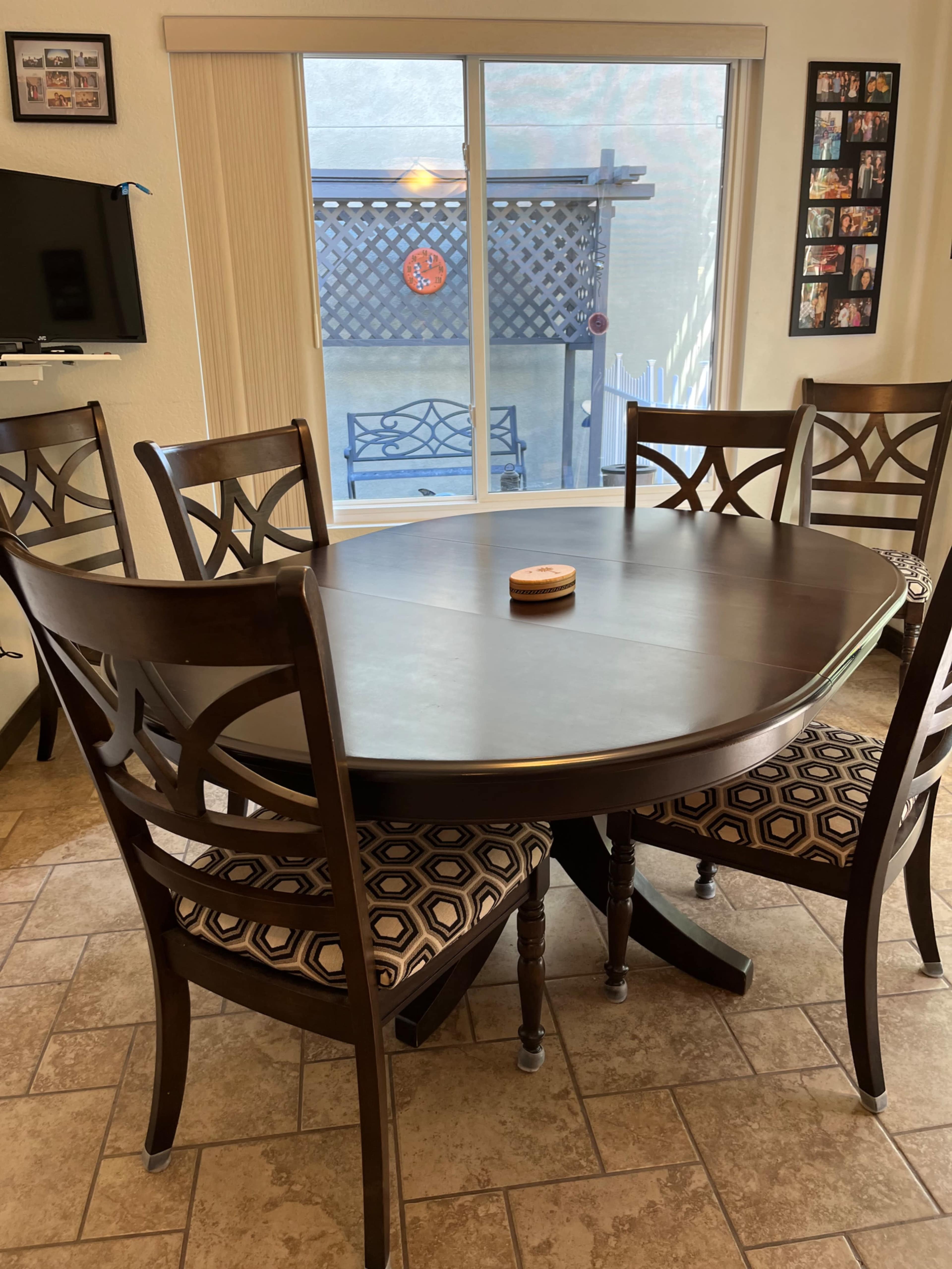 A round wooden dining table surrounded by four chairs is positioned in a brightly lit room with a window overlooking a patio.