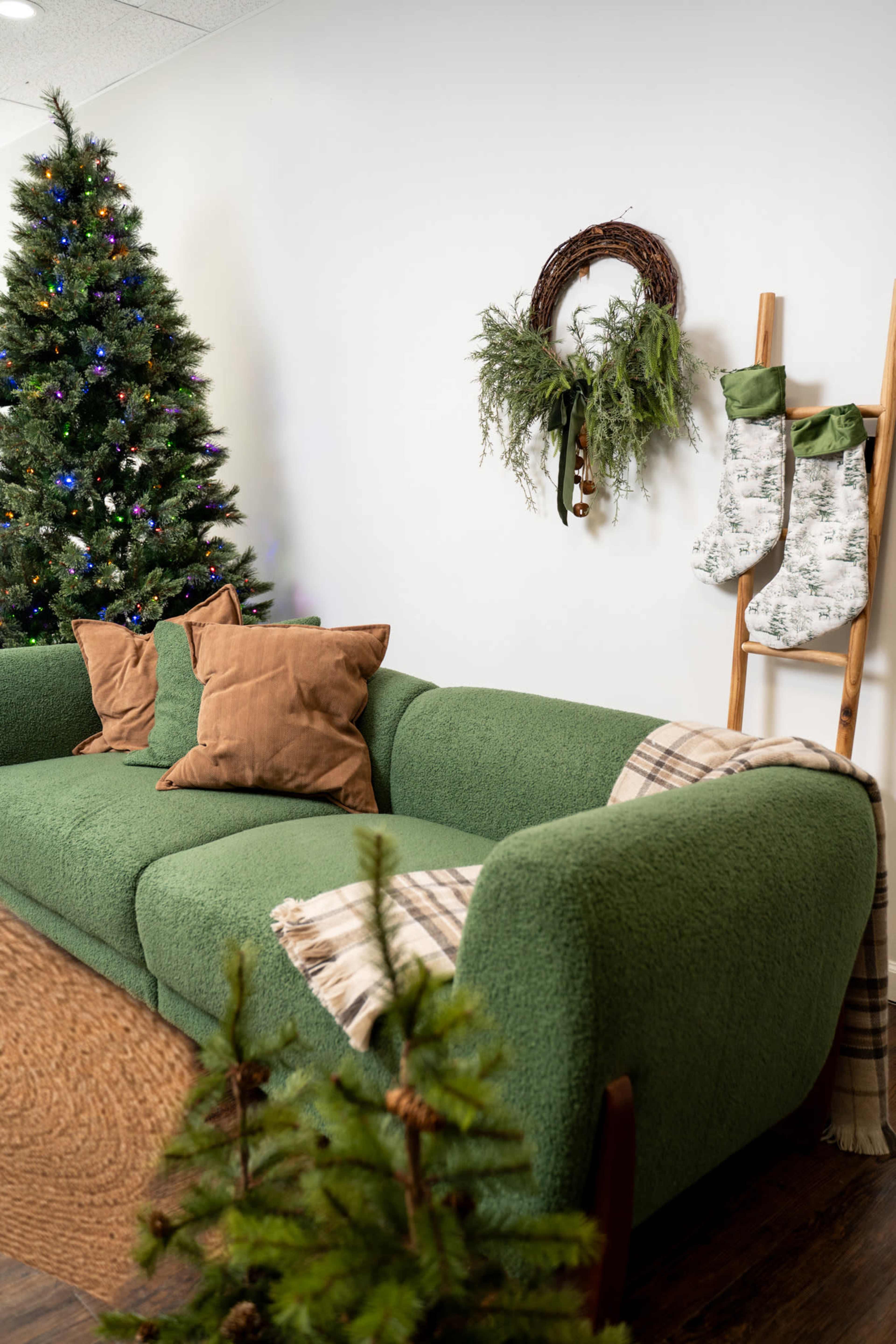 The image shows a cozy living room scene featuring a green sofa, decorated Christmas tree, a wreath on the wall, and festive stockings hung on a ladder.