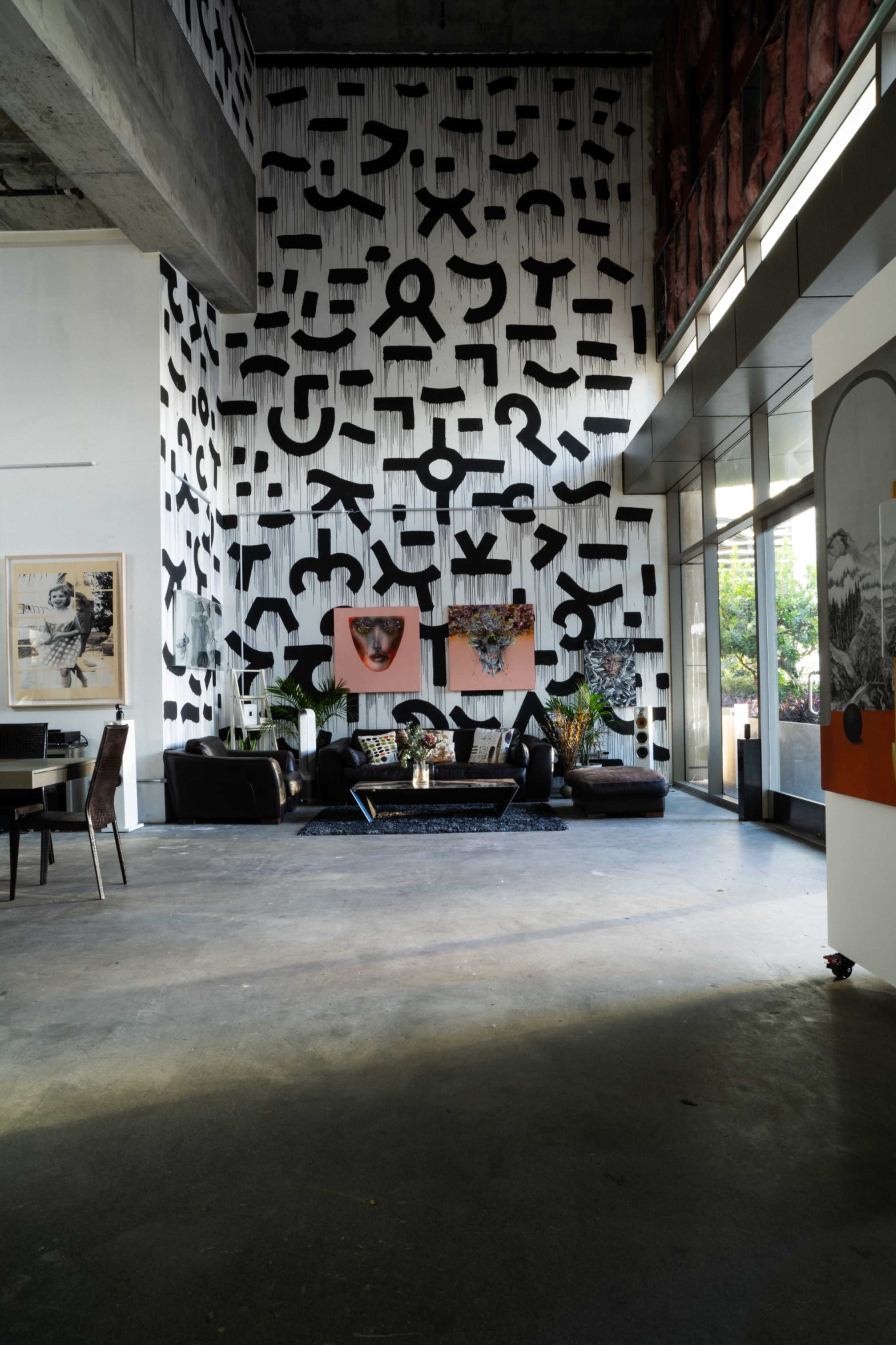 The image features a modern interior space with a high wall adorned with black abstract patterns, complemented by a dark sofa, plants, and artwork in a contemporary setting.