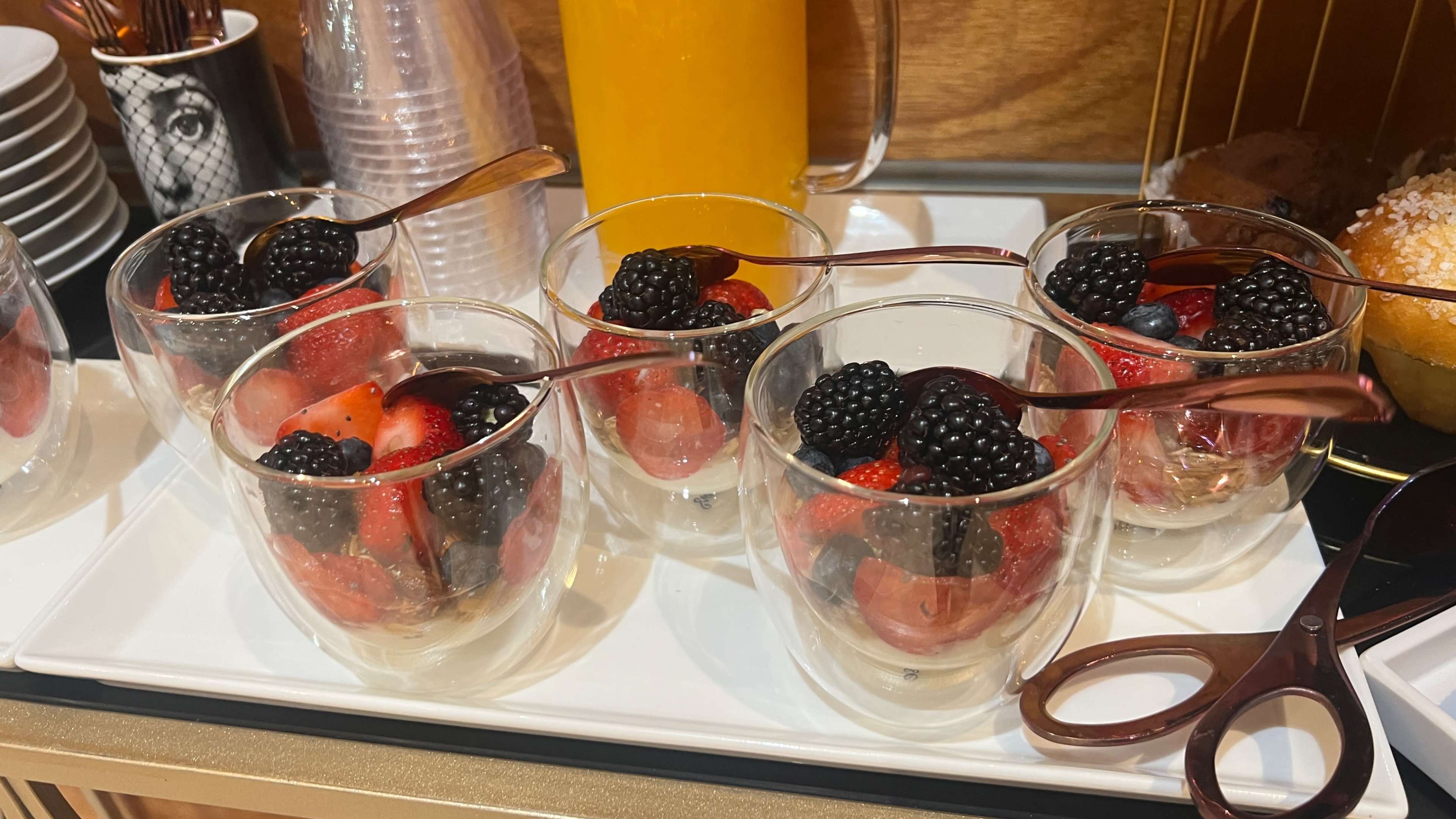 Five clear cups filled with mixed berries, including strawberries, blueberries, and blackberries, sit on a white serving tray beside a pitcher of orange juice.