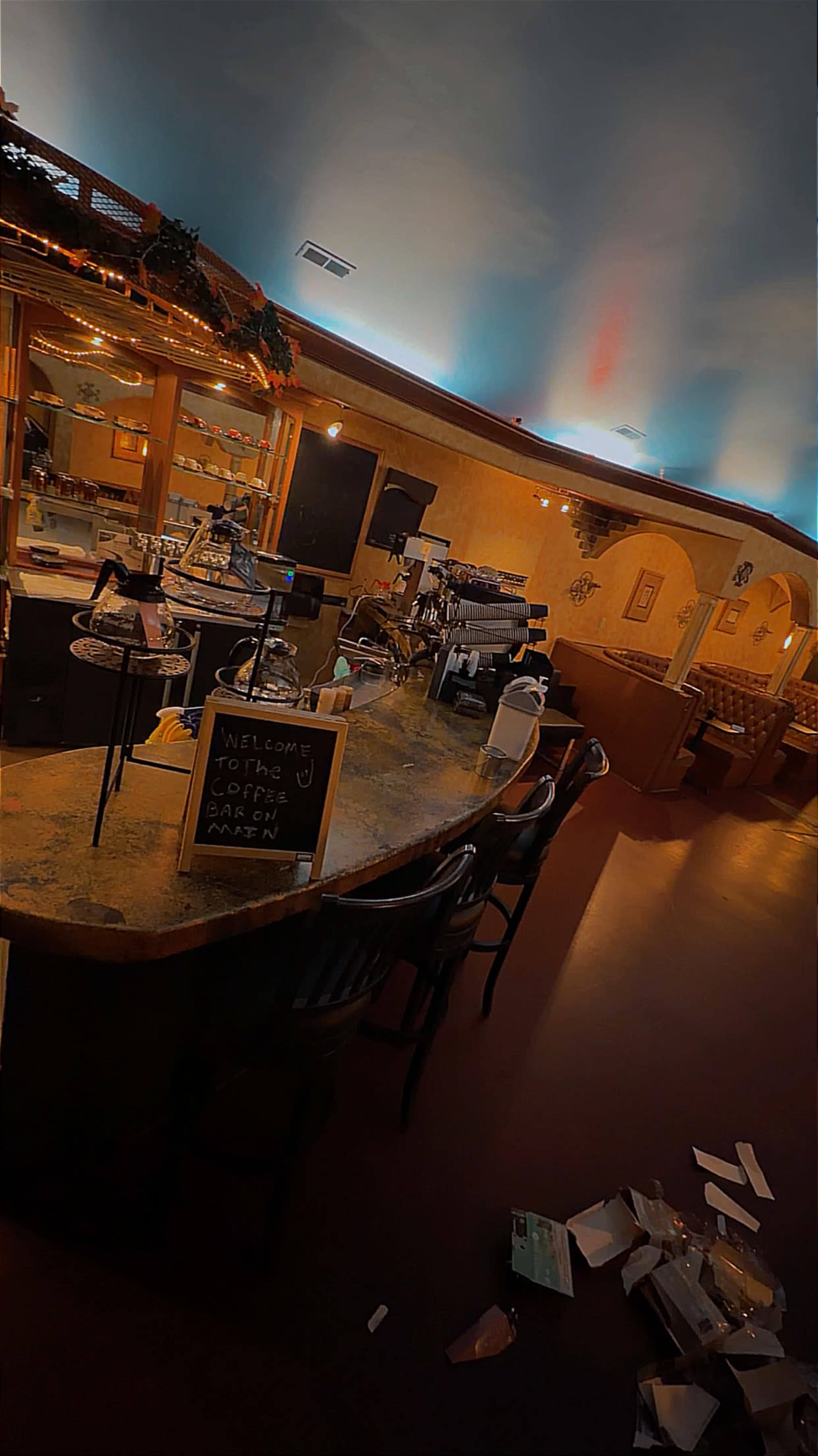 The image shows a bar area with a countertop, a sign welcoming patrons, and empty tables in a restaurant setting.