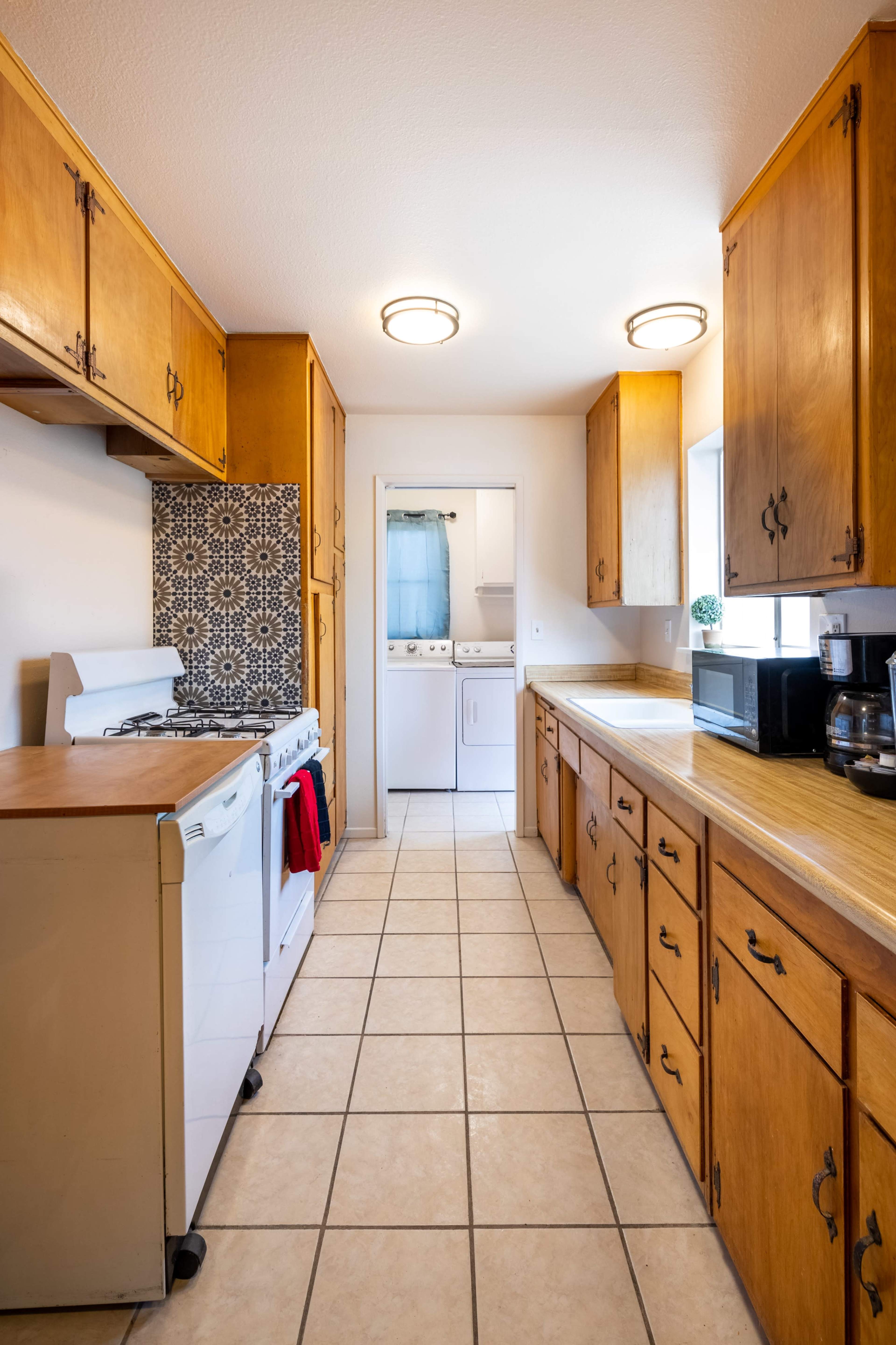 A narrow kitchen features wooden cabinets, a gas stove with a red towel, and a laundry area visible through a door at the end.
