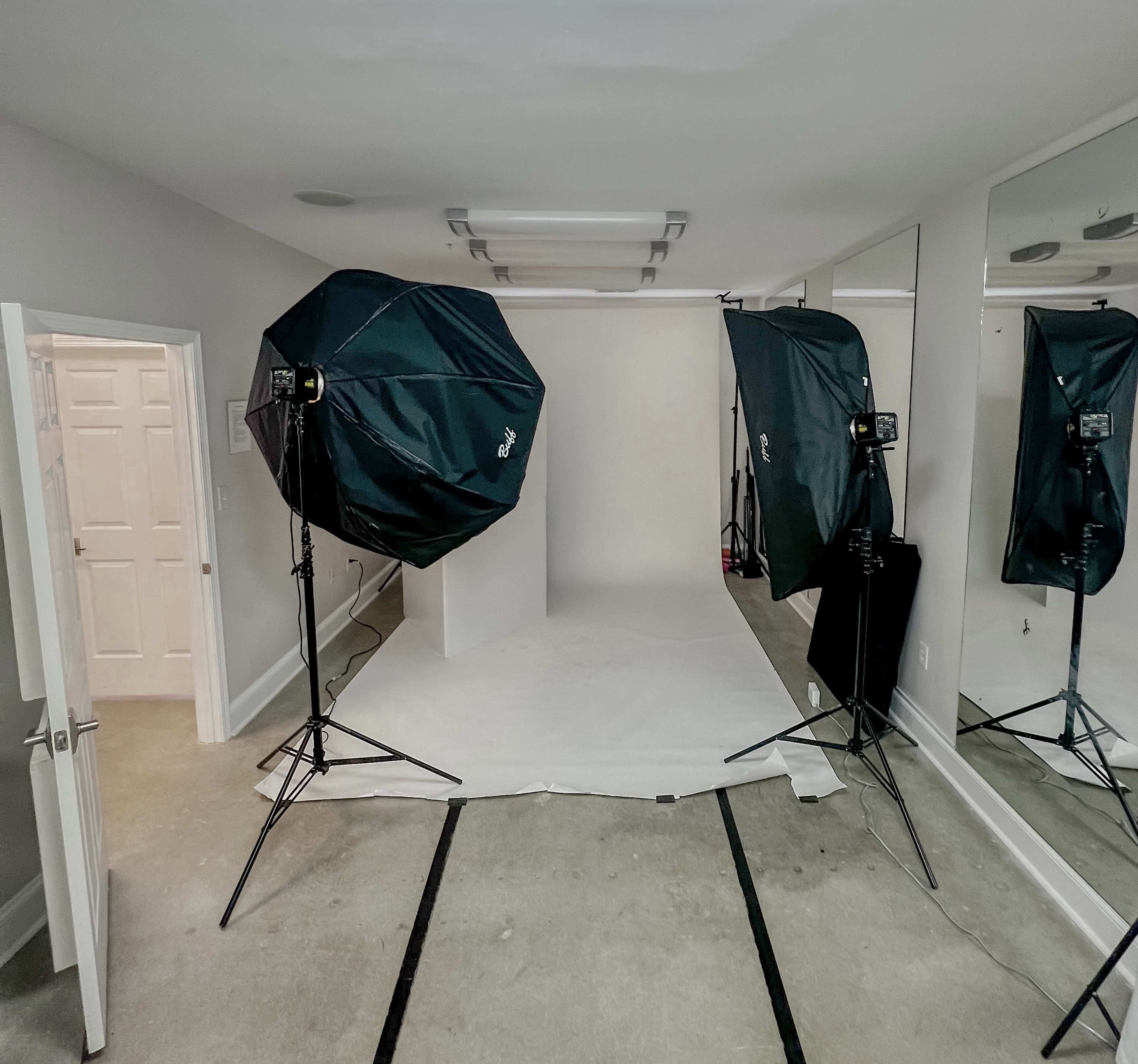 A photography studio equipped with two large softlight umbrellas facing a seamless backdrop, with mirrors and a white door visible in the background.