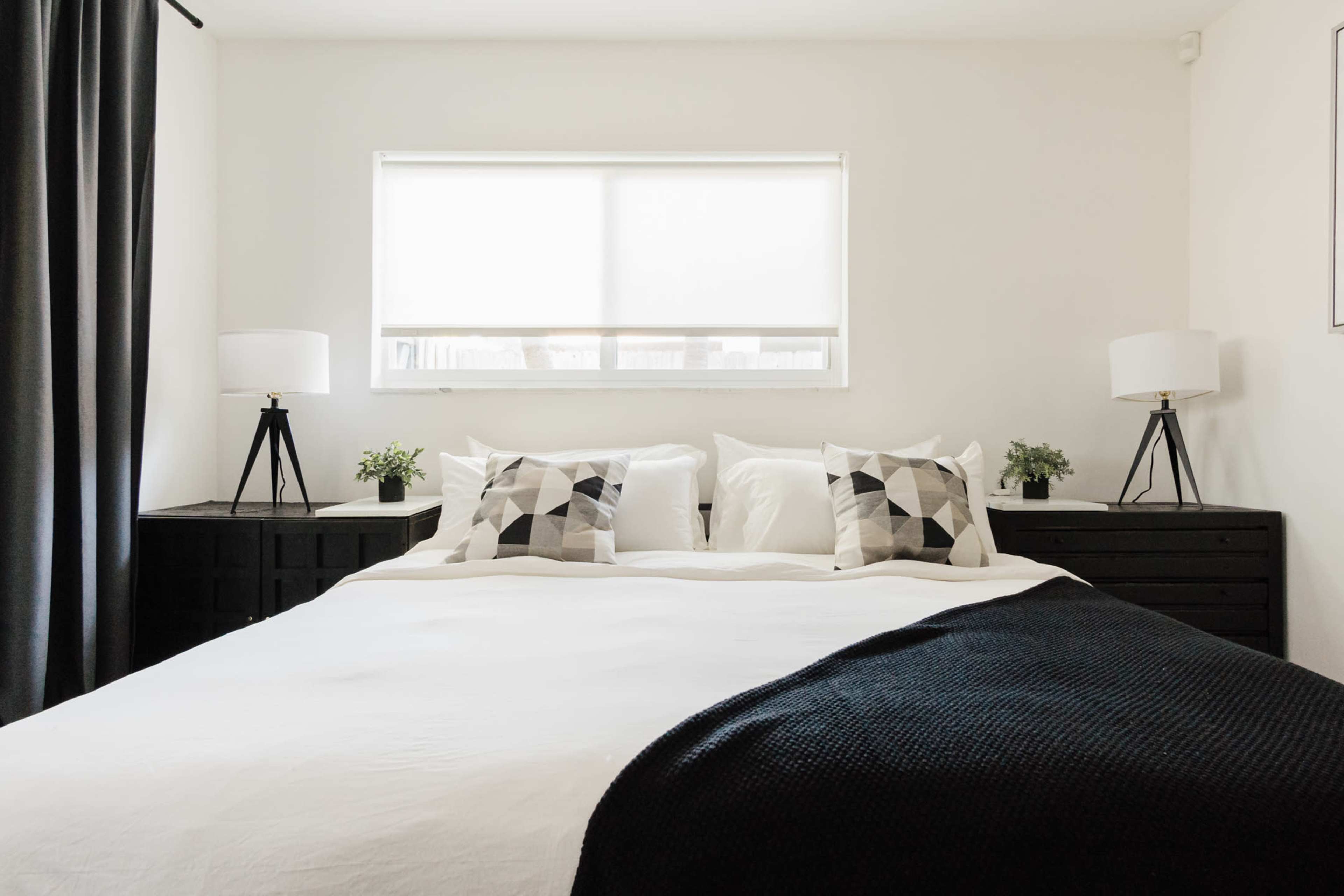 A neatly arranged bedroom features a bed with white linens, two patterned pillows, and nightstands on either side with lamps and small plants.