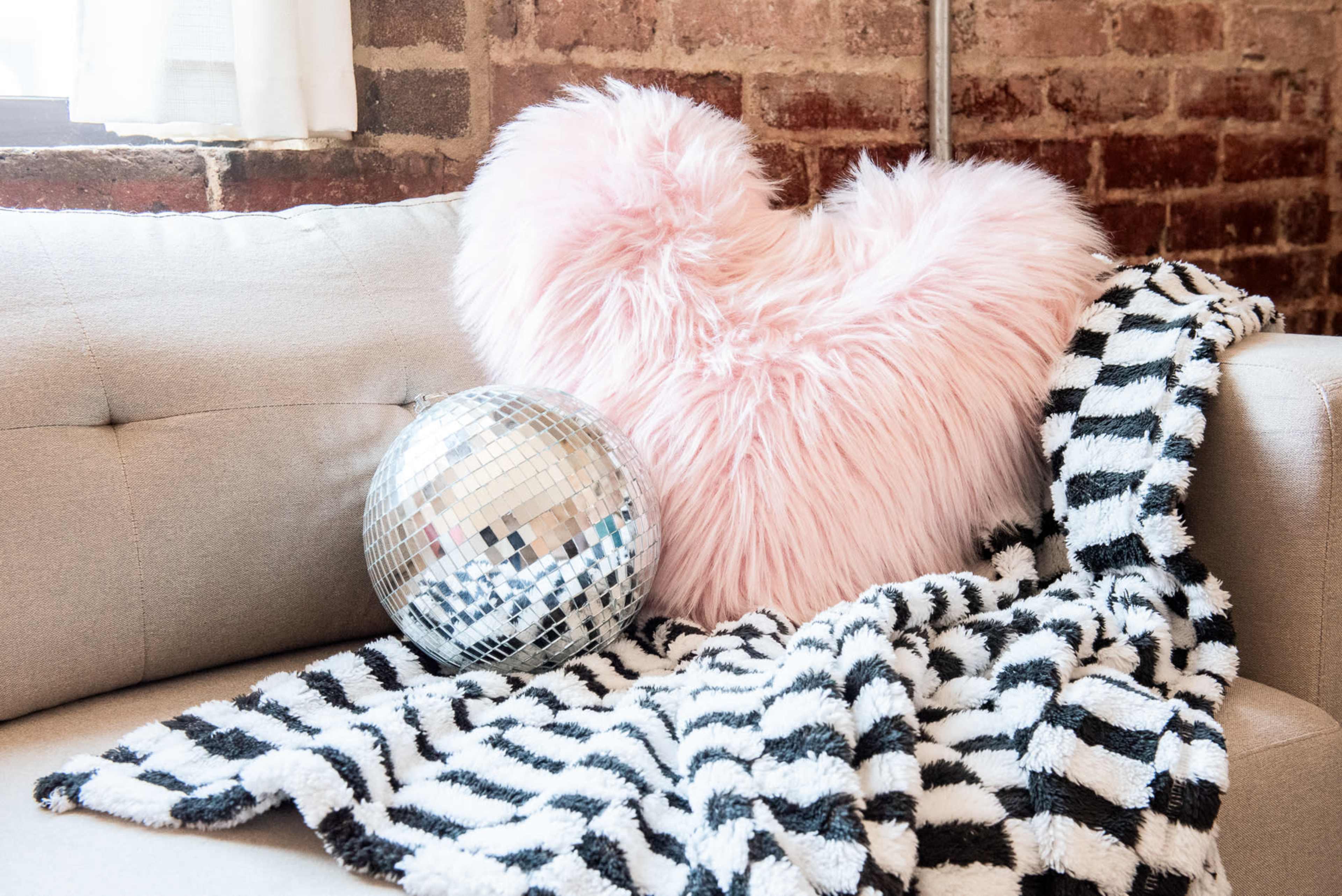 A pink heart-shaped pillow rests on a beige couch beside a silver disco ball and a black-and-white striped blanket.