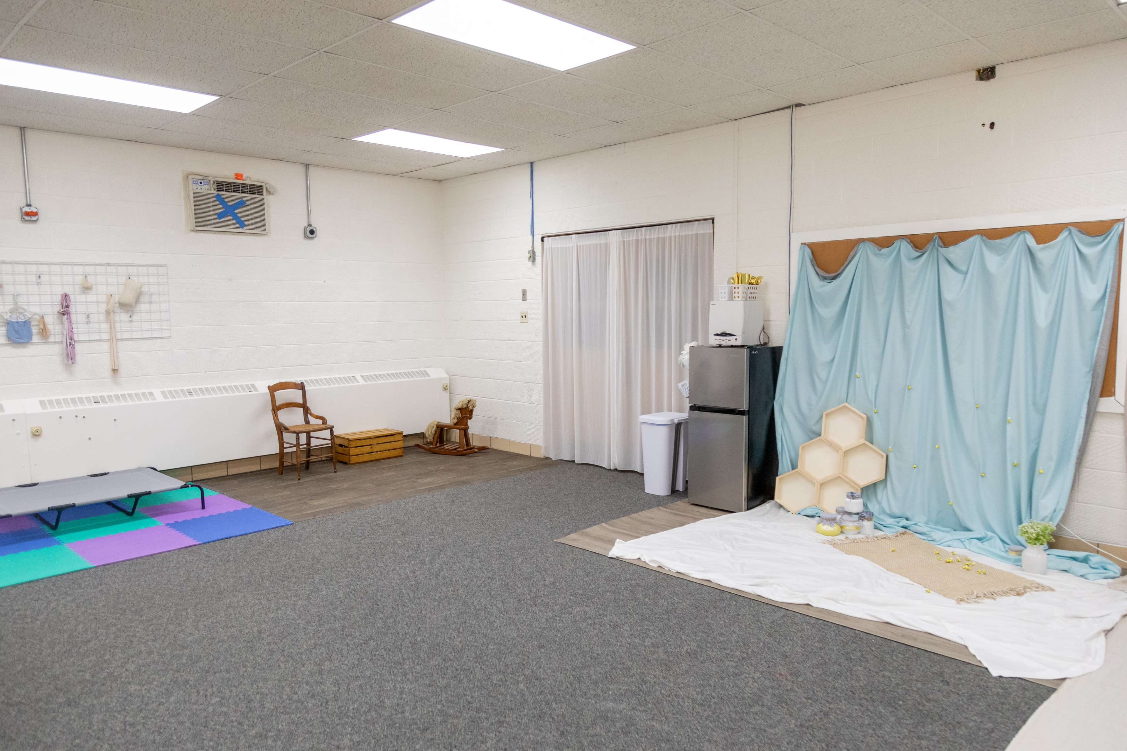 The image shows a spacious, empty room featuring a seating area with a wooden chair, a curtain-covered area, and a small display setup with a light blue backdrop and decorative elements on a white cloth.