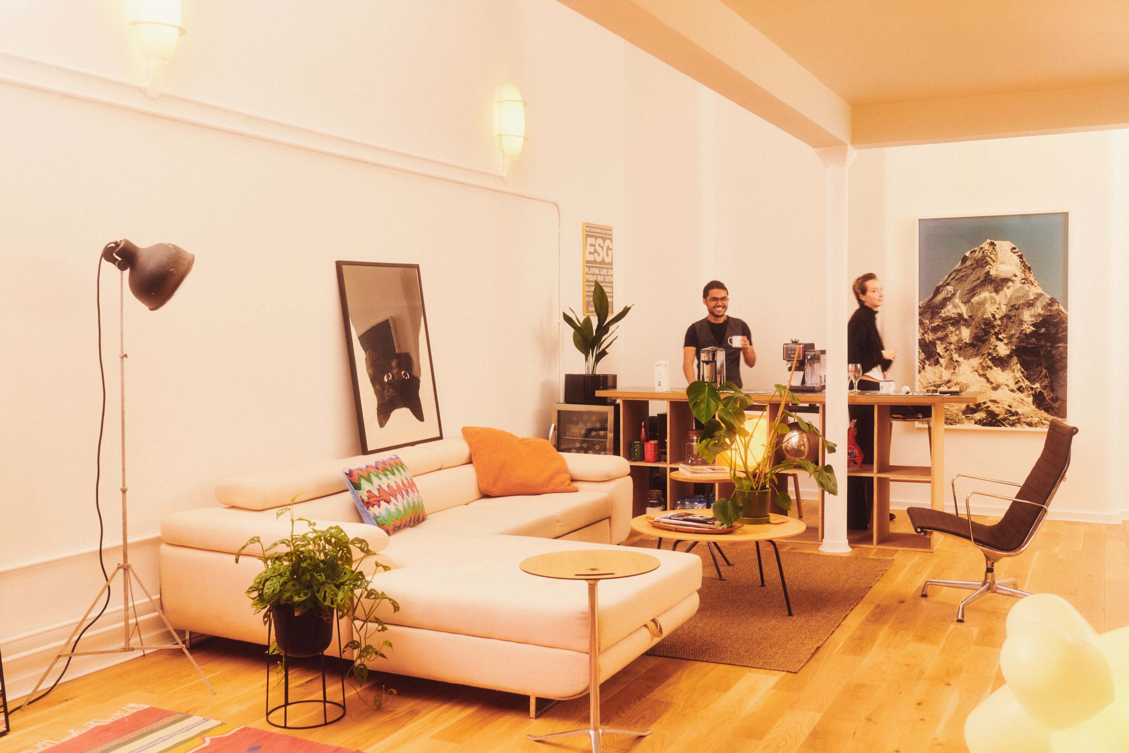 The image shows a modern office space featuring a light-colored couch, a wooden desk with two people, and a large mountain artwork on the wall.