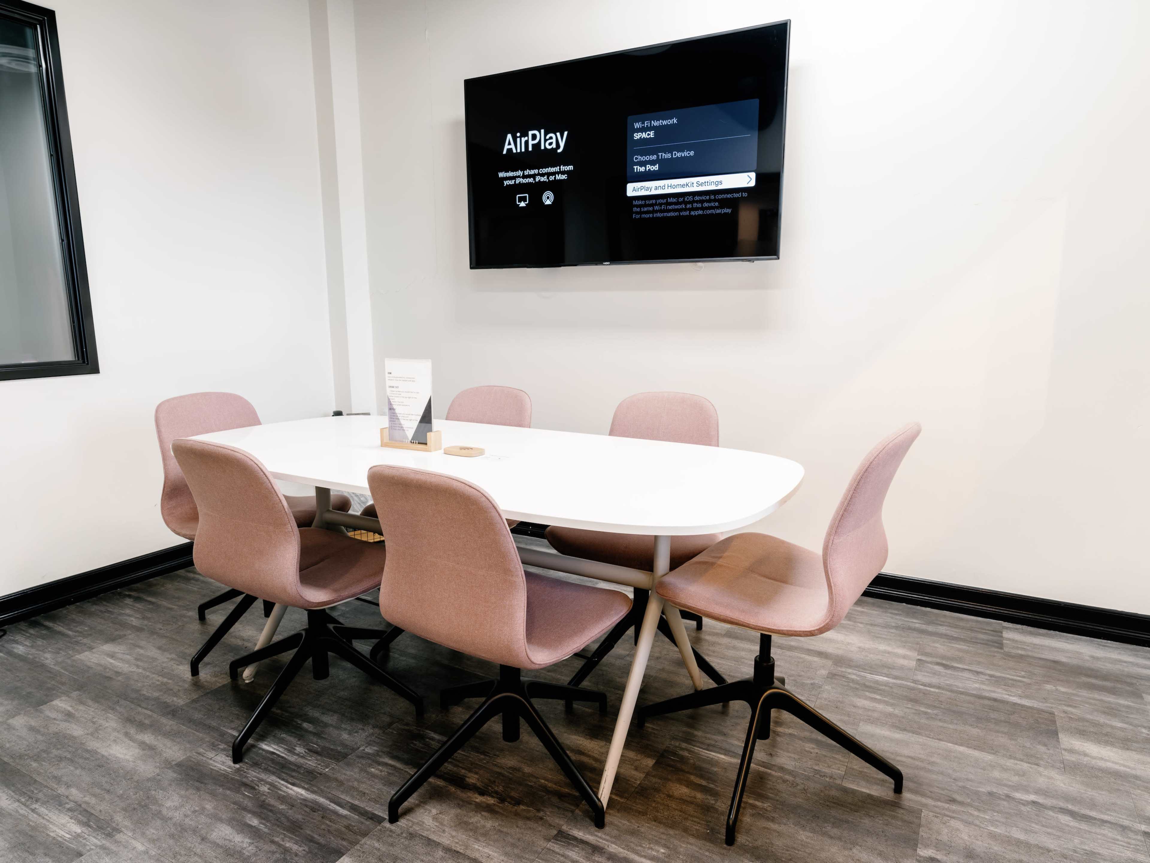 A modern conference room features a white oval table surrounded by six pink chairs, with a wall-mounted screen displaying an AirPlay menu.