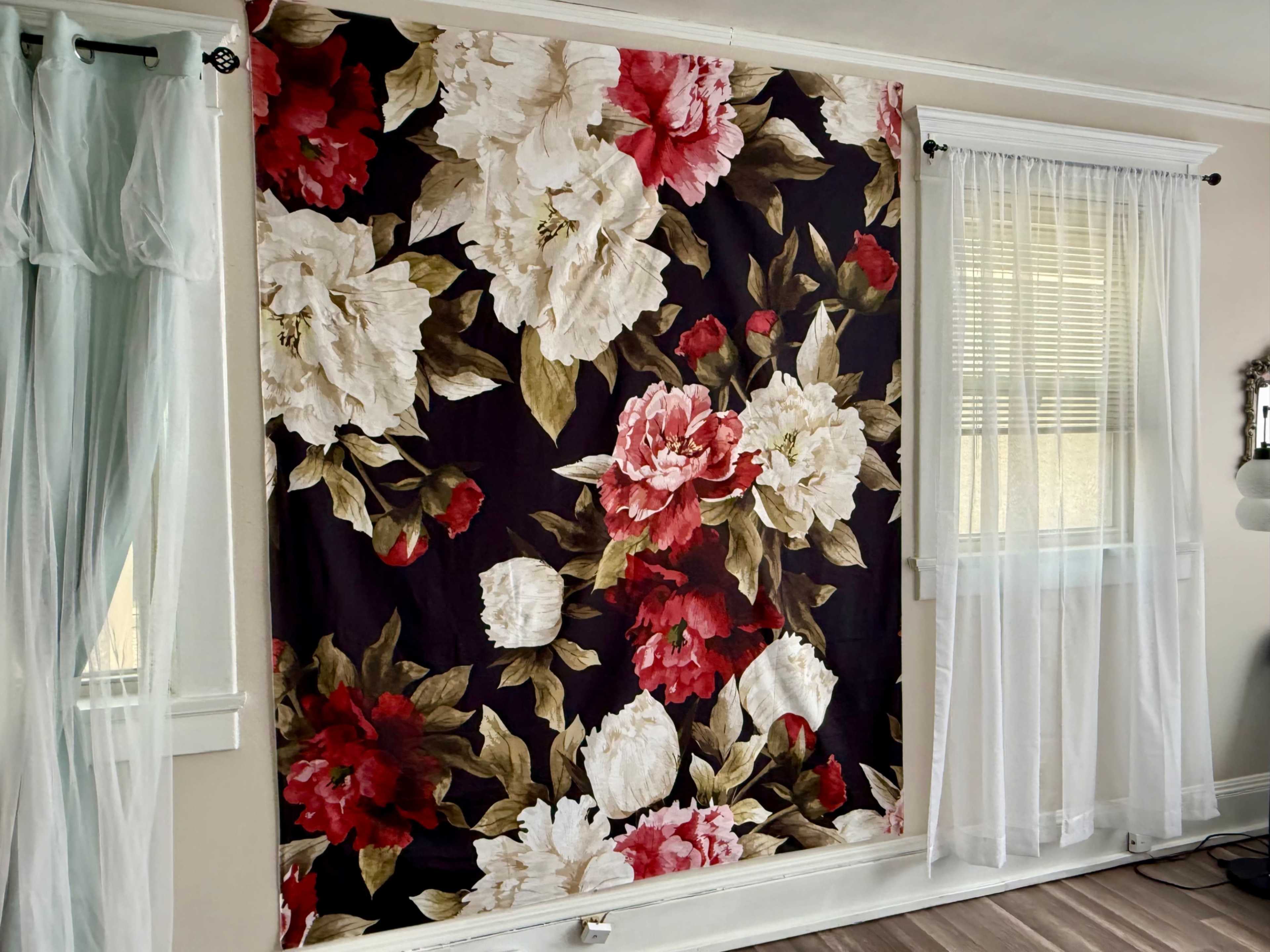 A large floral fabric panel in shades of red, white, and cream is displayed on a wall beside sheer curtains in a bright room.