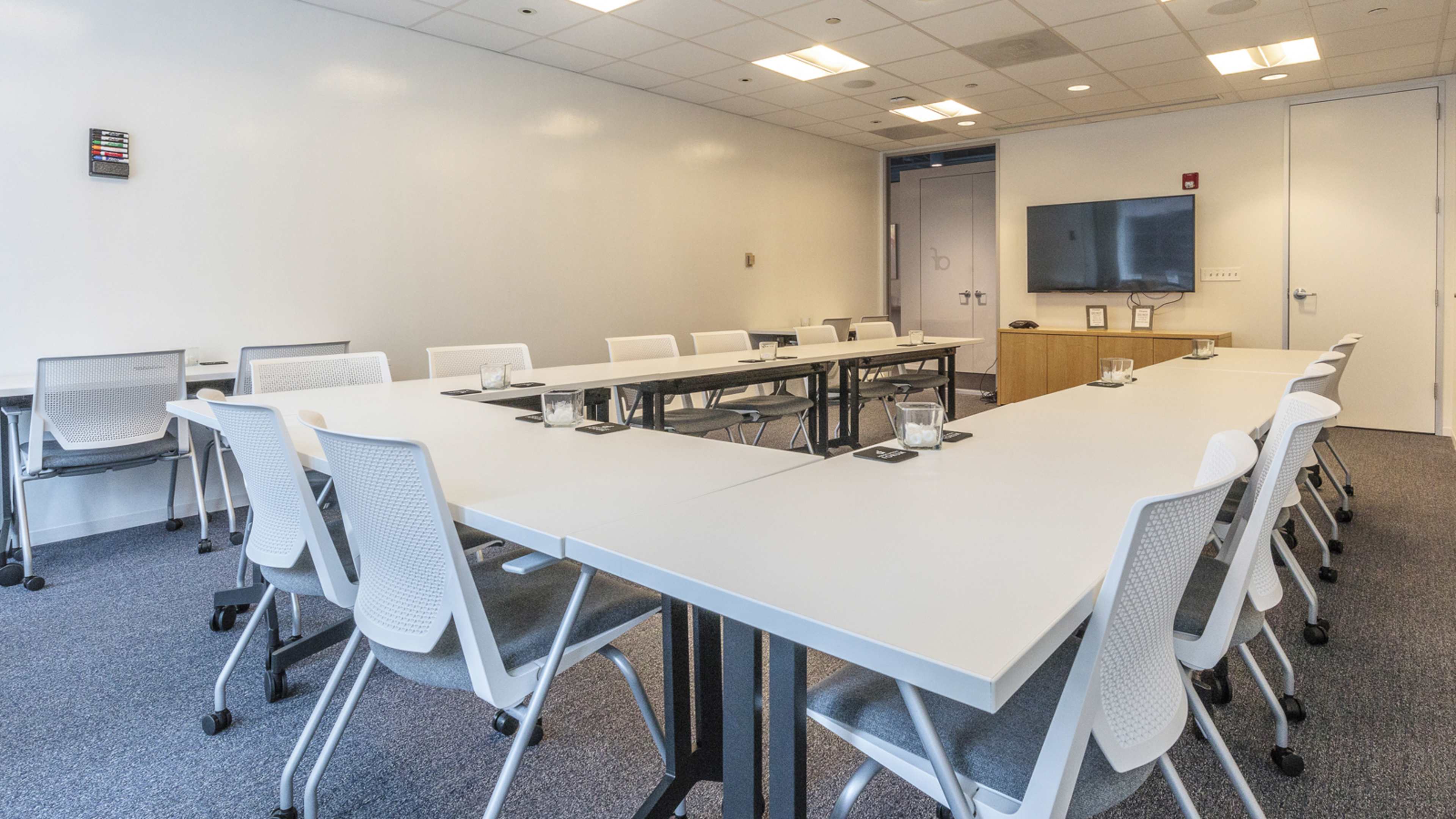 A modern conference room features multiple tables arranged in a U-shape, with chairs, a television mounted on the wall, and a glass panel door.