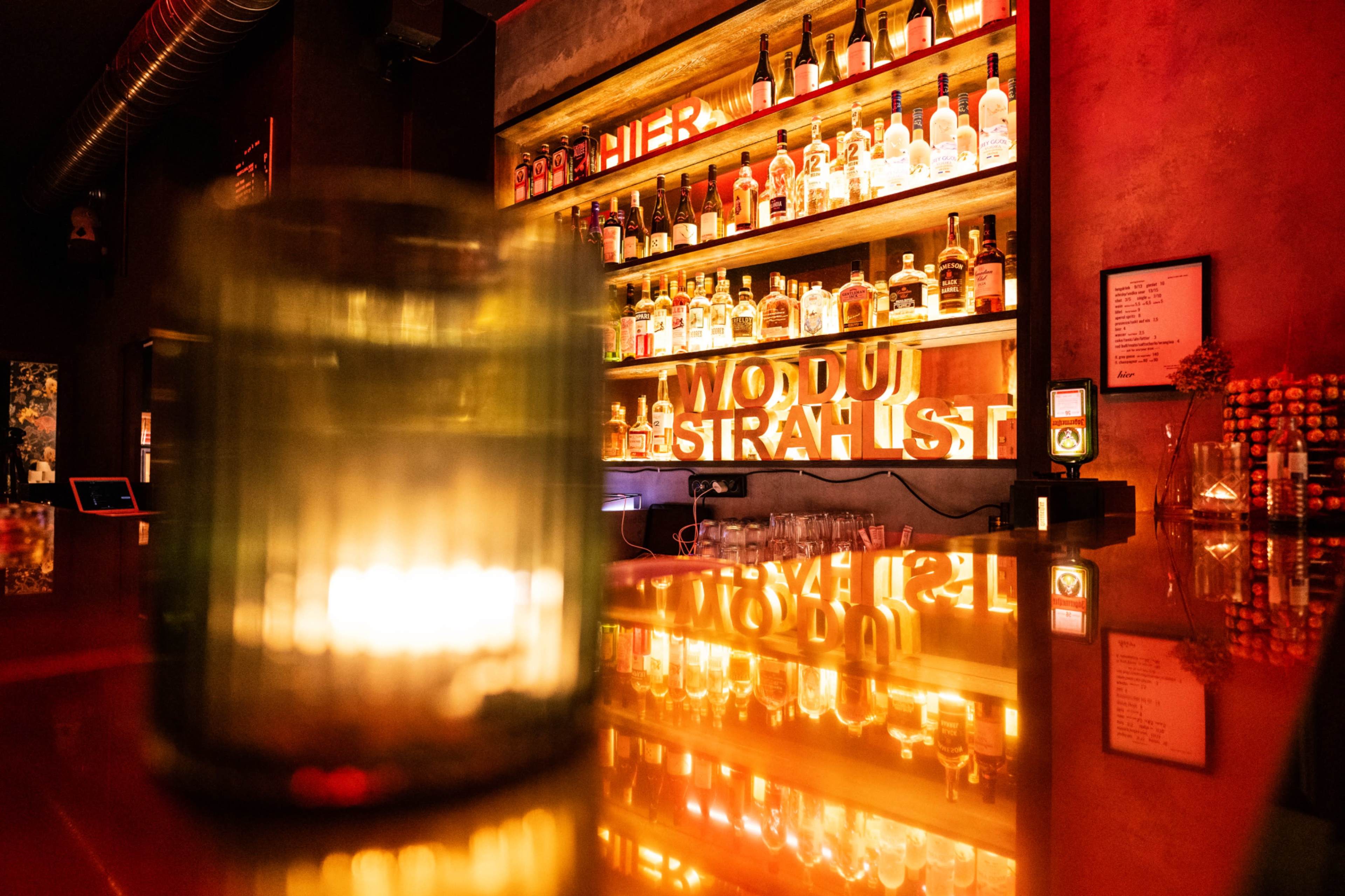 A well-lit bar displays a variety of liquor bottles on shelves against a warmly illuminated backdrop, with a candle on the counter in the foreground.